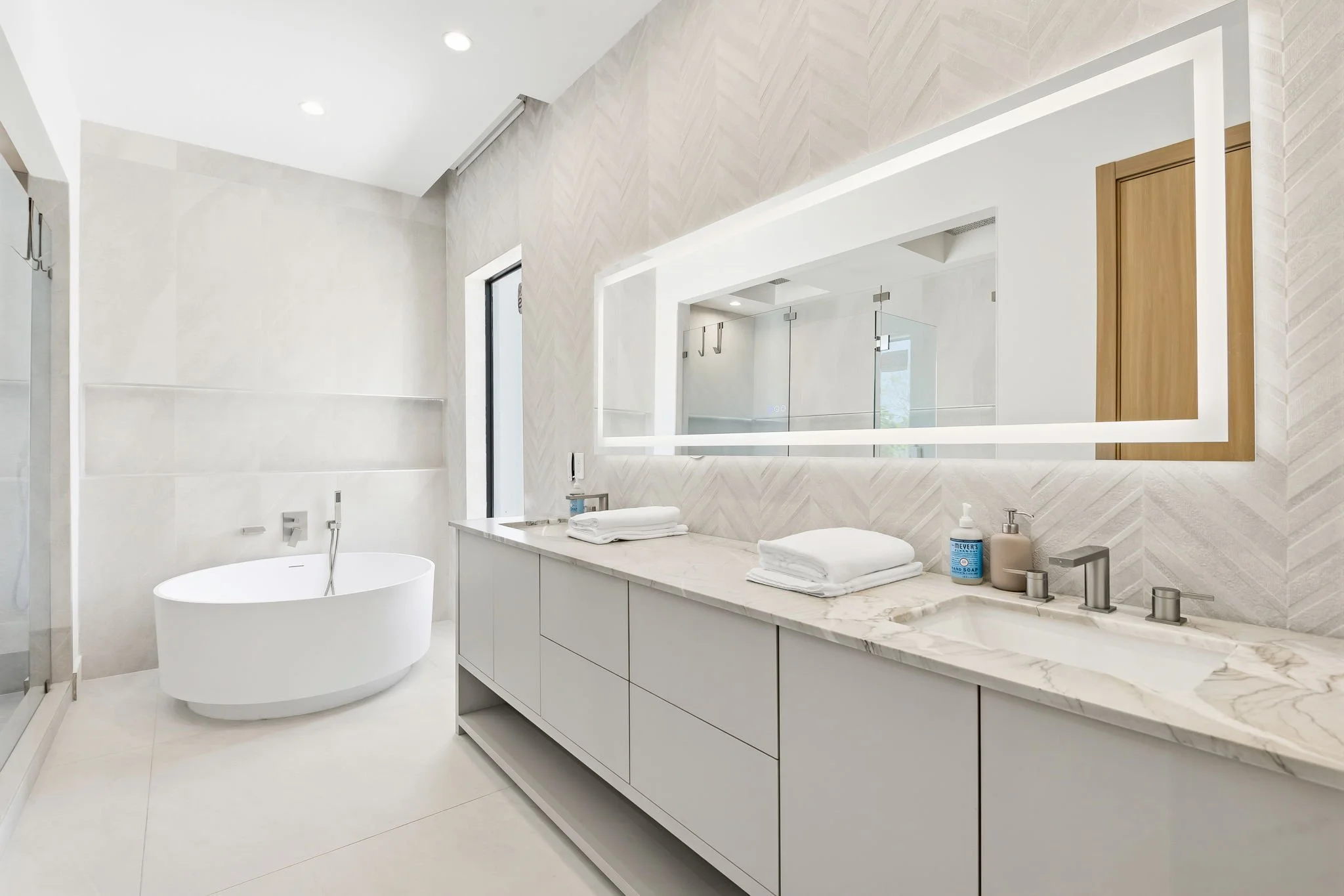Modern bathroom with a white freestanding bathtub, a marble countertop with folded towels, soap, and hand sanitizer, large mirror, wooden door, and clean minimalist design.