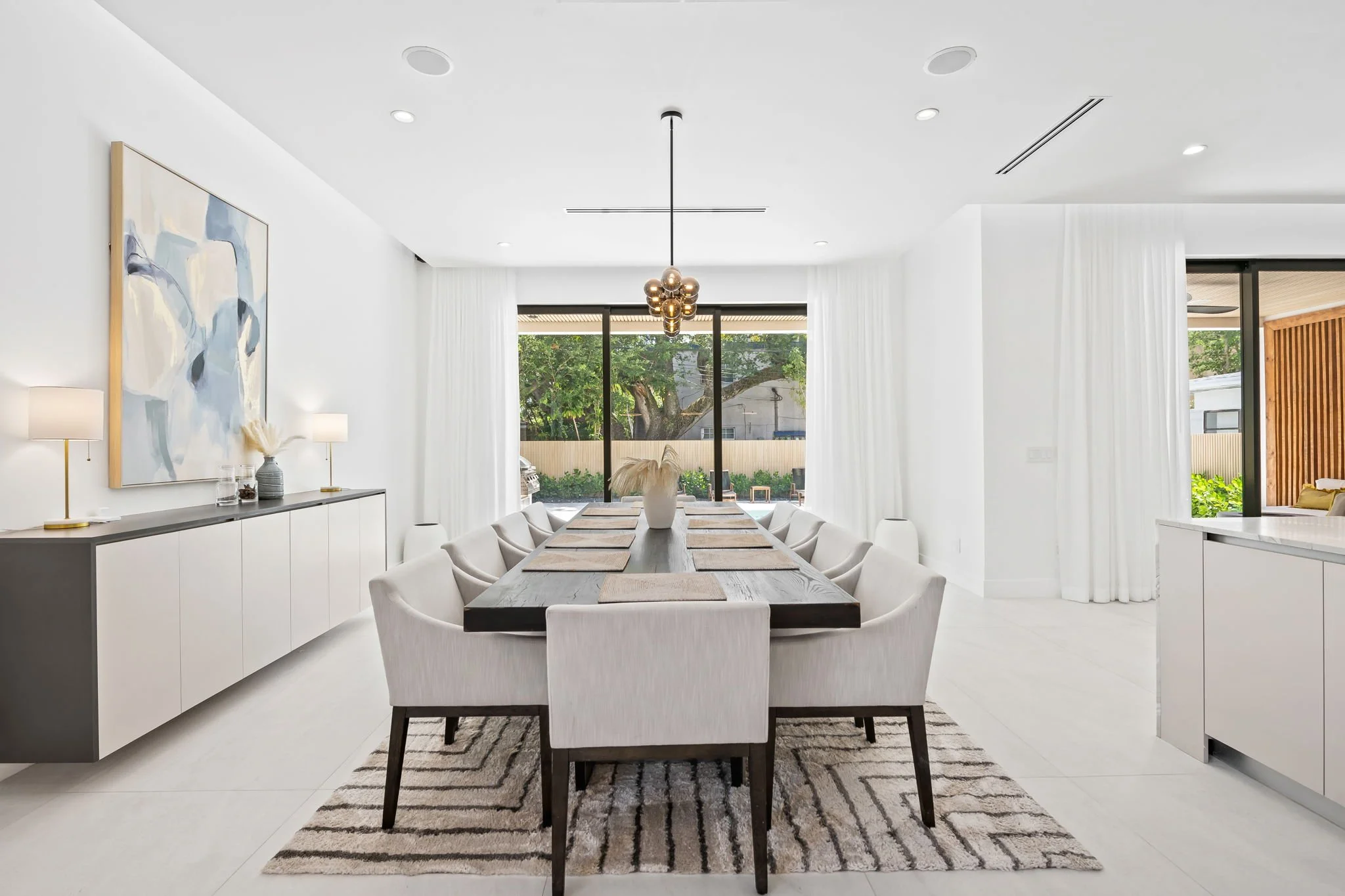 Modern dining room with a large wooden table, white upholstered chairs, a striped area rug, and a contemporary chandelier. Large windows with sheer curtains overlook a backyard with trees.