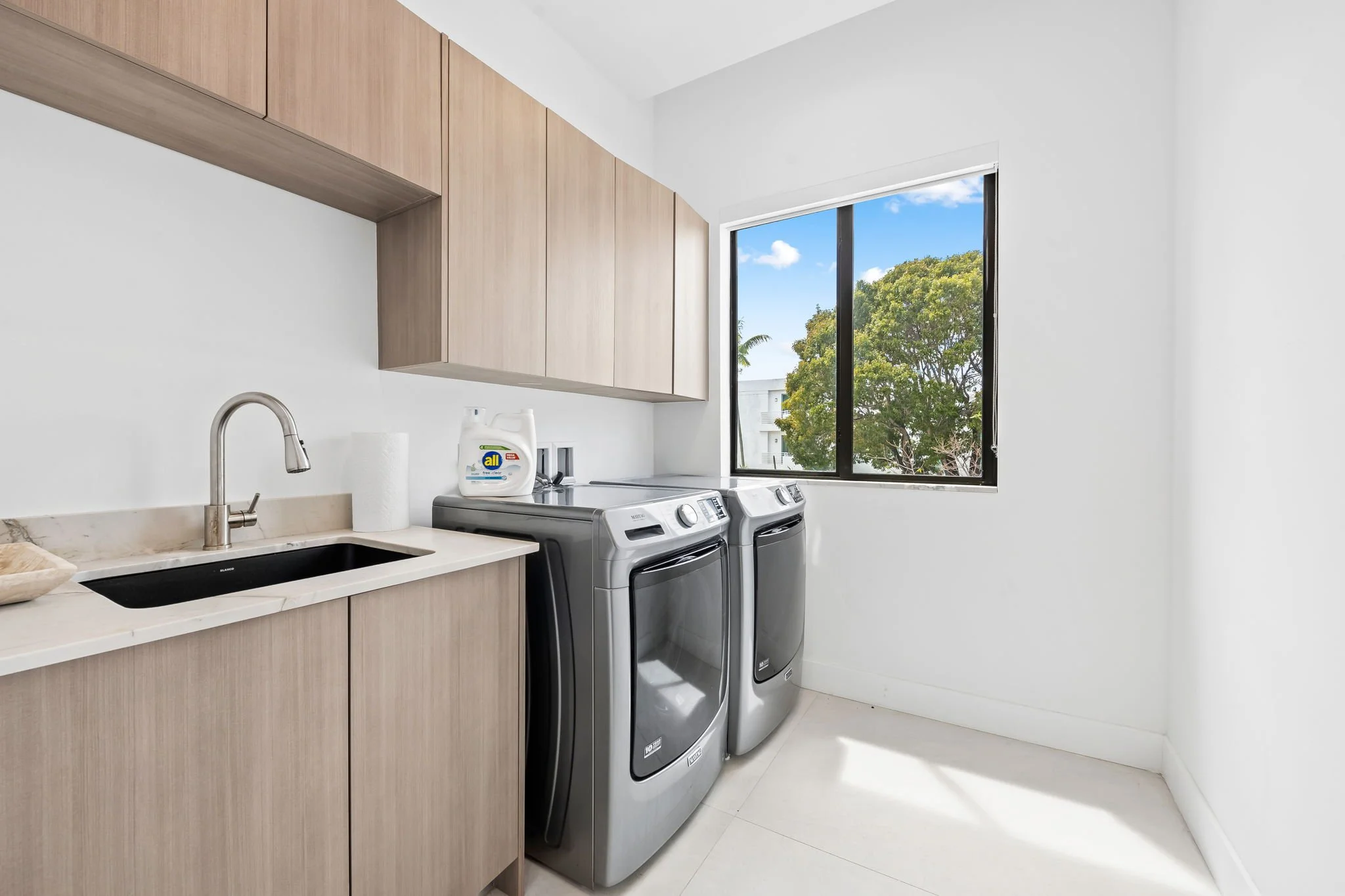 A laundry room with light wood cabinets, a white marble countertop, a stainless steel sink, a bottle of All laundry detergent, and a window showing trees and blue sky.