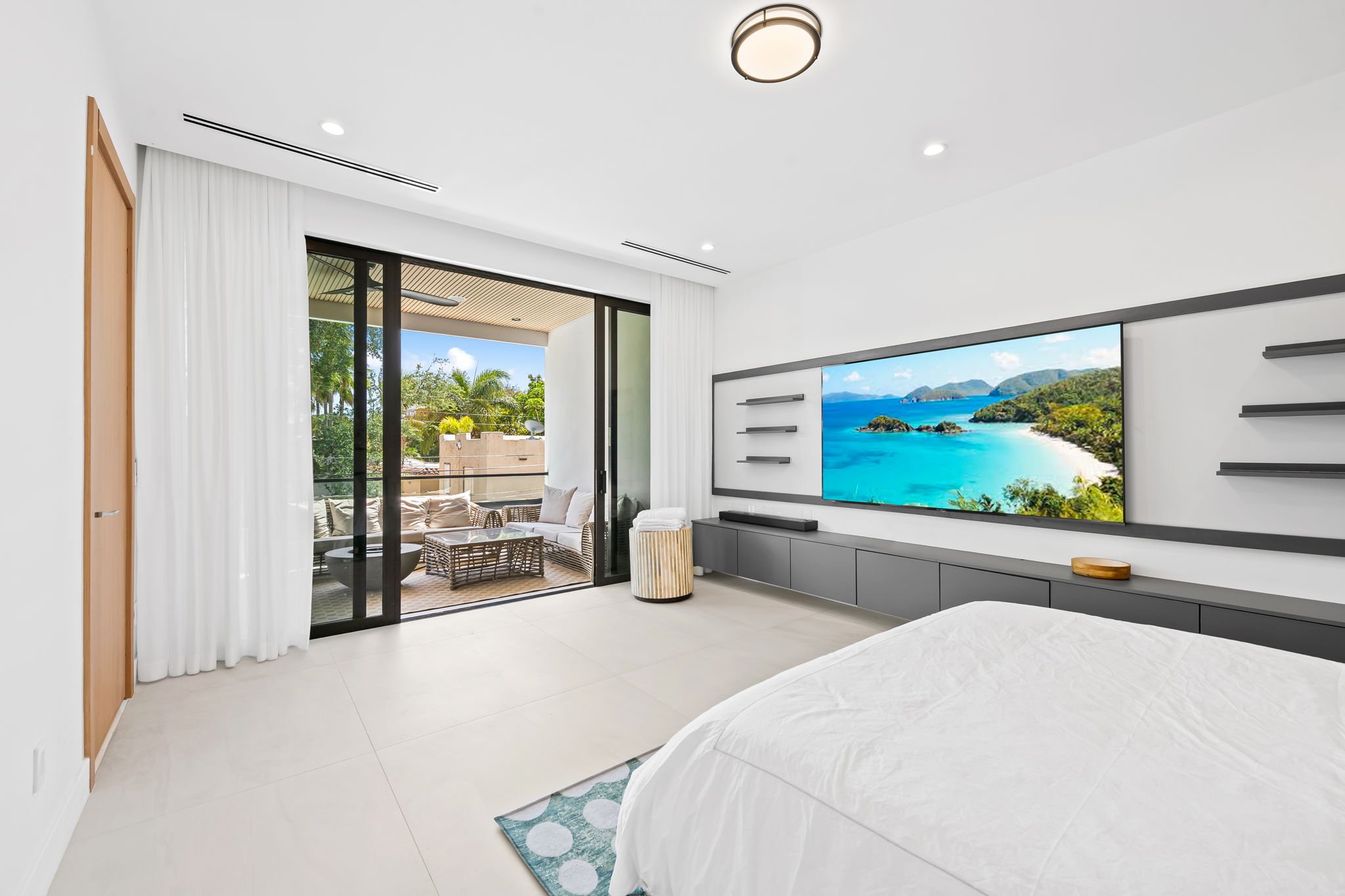 Modern living room with slide doors opening to a balcony with outdoor furniture, overlooking greenery and ocean view, with white walls, a large TV, shelves, and minimalist decor.
