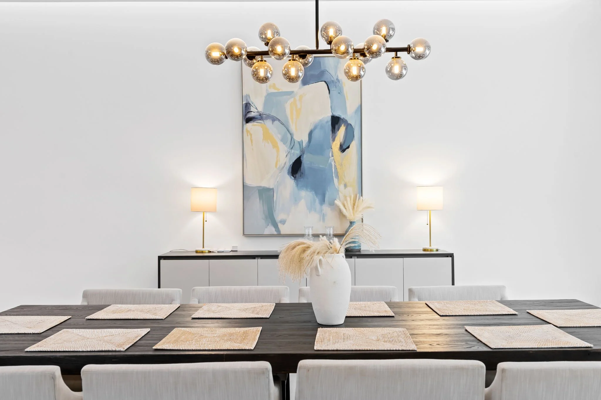 Modern dining room with a dark wood table, white chairs, beige placemats, a white vase with pampas grass, two wall lamps, an abstract painting, and a contemporary chandelier with multiple glass globes.