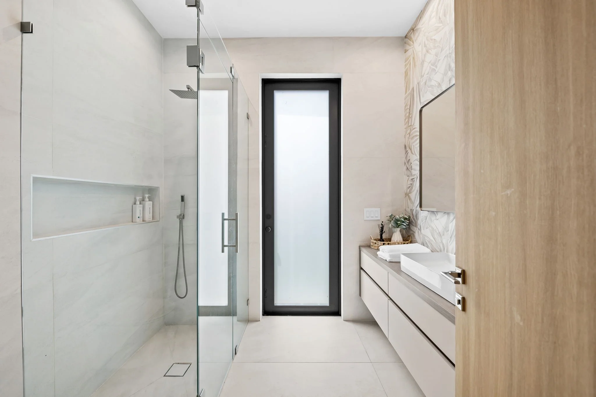 Modern minimalist bathroom with a walk-in shower, black-framed frosted glass door, white vanity with a vessel sink, and decorative tiles on one wall.