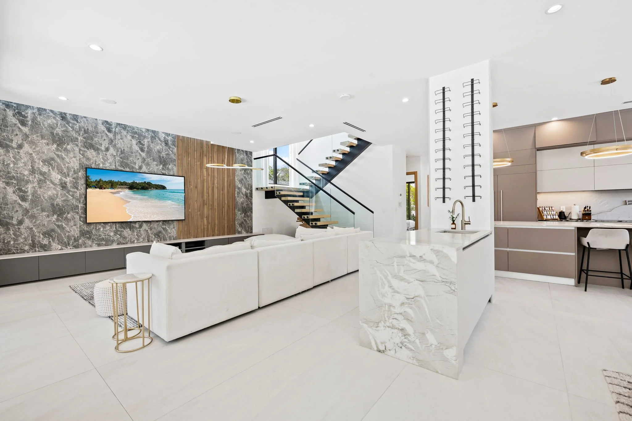 Modern open-plan living room and kitchen with white walls, marble and wood accents, a staircase with glass railing, and a wall-mounted TV displaying a beach scene.