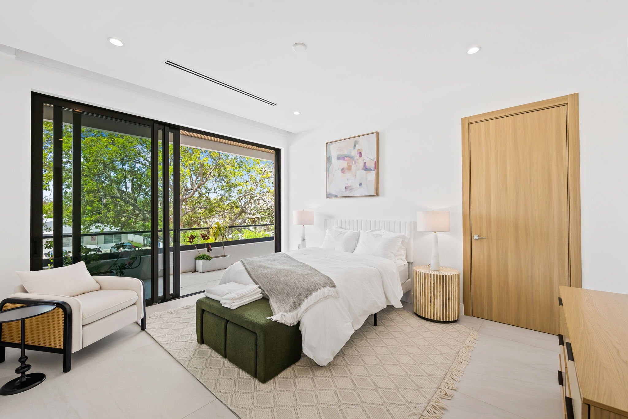 Modern bedroom with large window showing green trees, white walls, bed with white linens, side table with lamp, framed abstract art, wooden closet door, and beige area rug.