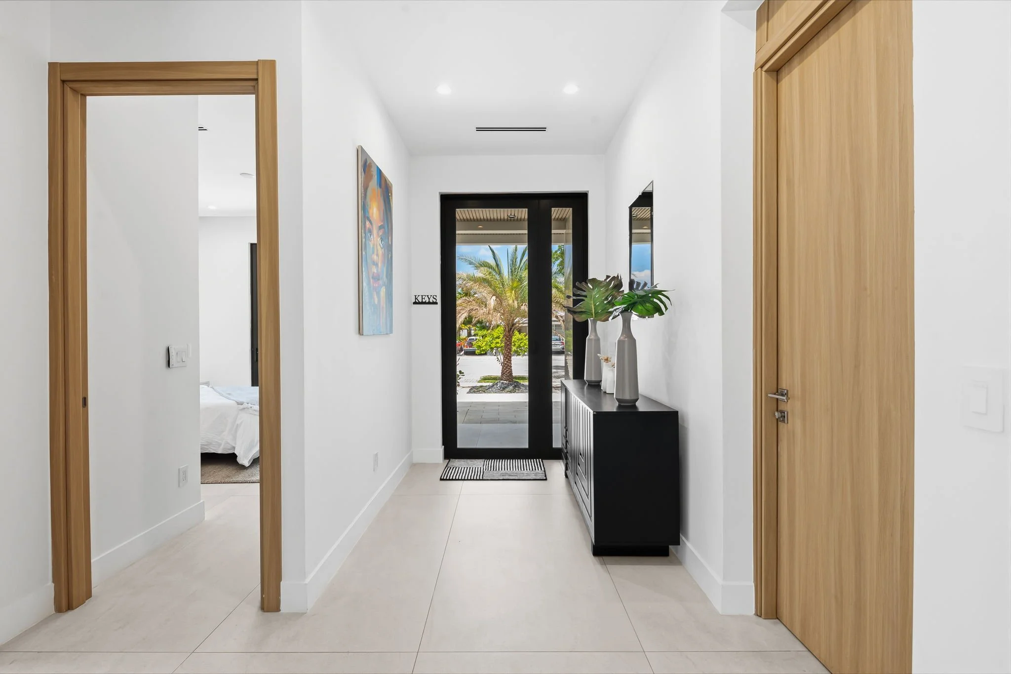 Modern entryway with white walls, tiled flooring, wooden door frames, a black door with glass panels showing an outdoor view of palm trees, a black sideboard with white vases and green plants, a colorful abstract painting, and a small sign that reads