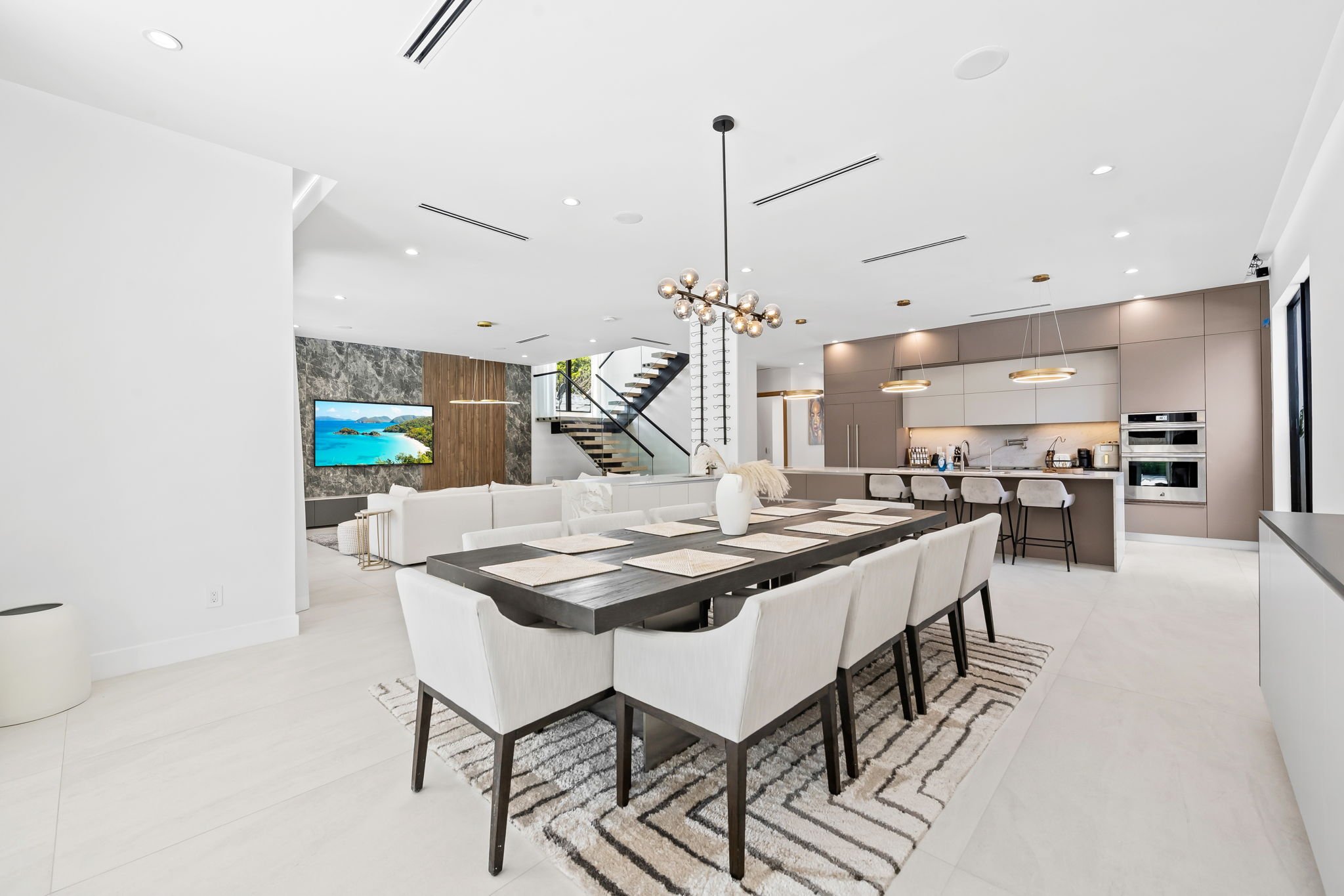 Open-concept modern living and kitchen area with white walls, a wooden accent wall, and a staircase with glass railing. The space features a dining table with white chairs, a large flat-screen TV, and contemporary lighting fixtures.