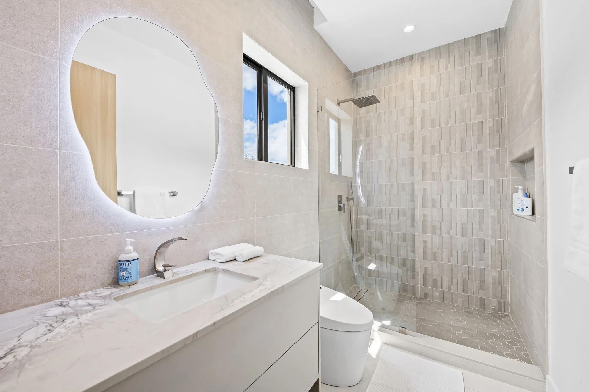 Modern bathroom with beige tiled walls, a marble countertop sink, a wall-mounted mirror, a window showing blue sky and clouds, a glass-enclosed shower with a rainfall showerhead, lotion bottles on a shelf, and a toilet.