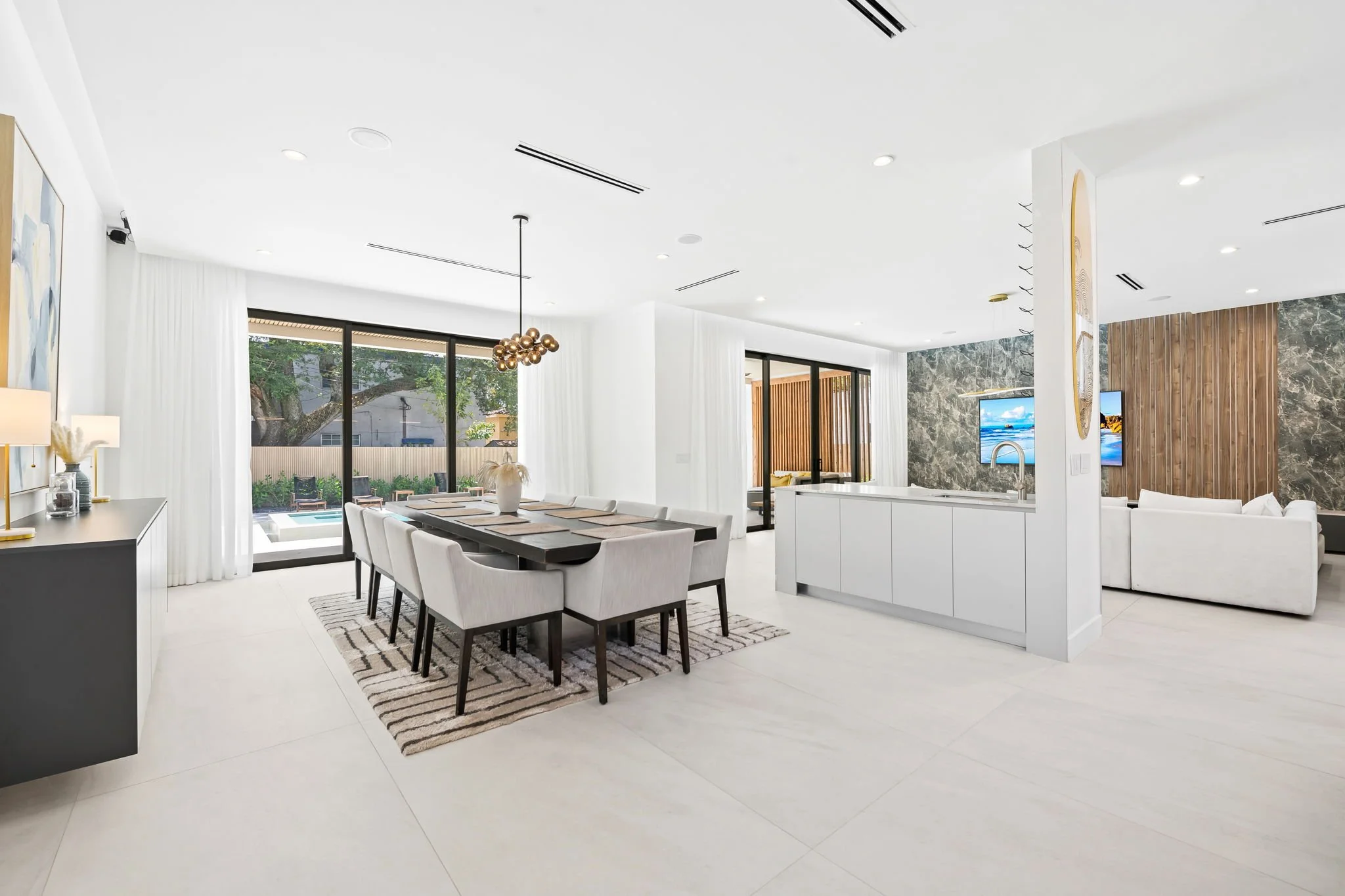 Modern interior living and dining room with white walls, large windows, black sliding doors, a dining table with eight chairs, a chandelier, and a living area with a white couch and TV.