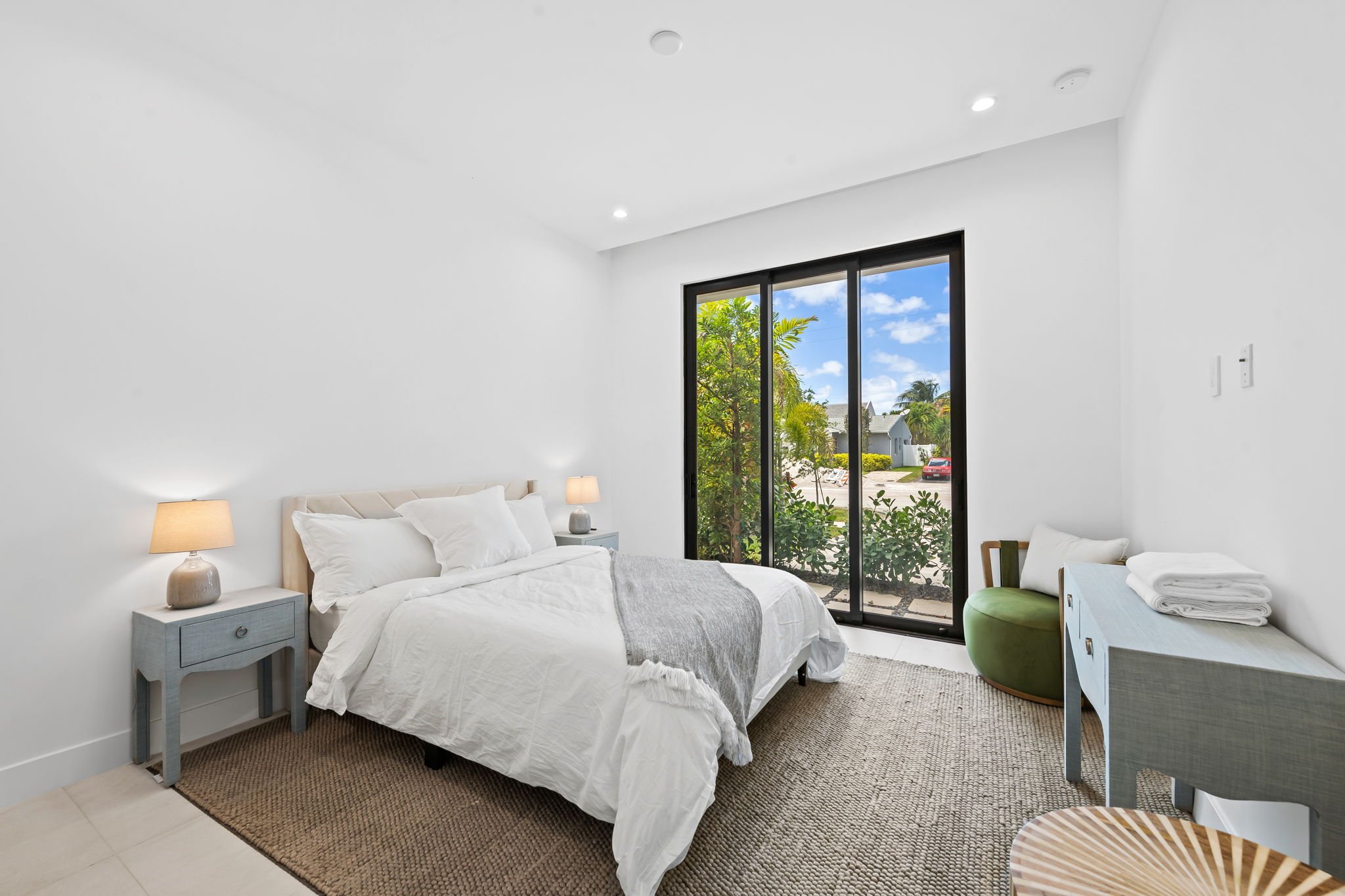A bright, modern bedroom with white walls and ceiling. It features a bed with white linens, a gray blanket, and white pillows, flanked by two nightstands with beige lamps. There is a framed sliding door with black trim leading outside, revealing lush