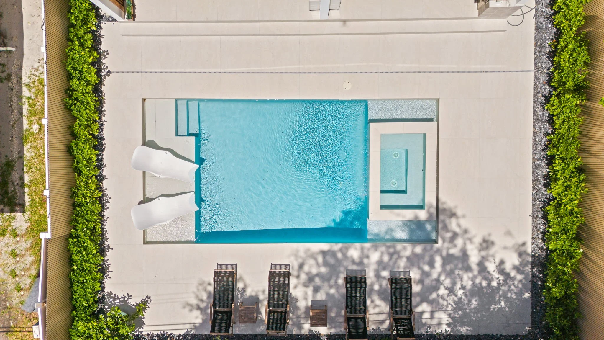 A backyard swimming pool with a white waterslide on the left side, surrounded by a concrete deck, with green shrubs and flowers along the top and right edges, and four chairs at the bottom of the image.