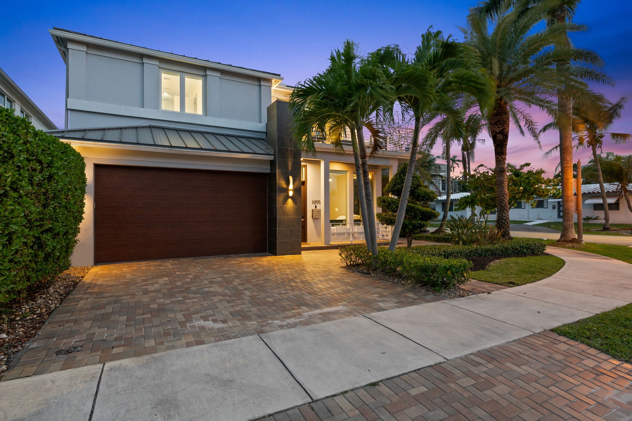 Modern two-story house with white exterior, large windows, a garage door, and tropical palm trees in the front yard during sunset.