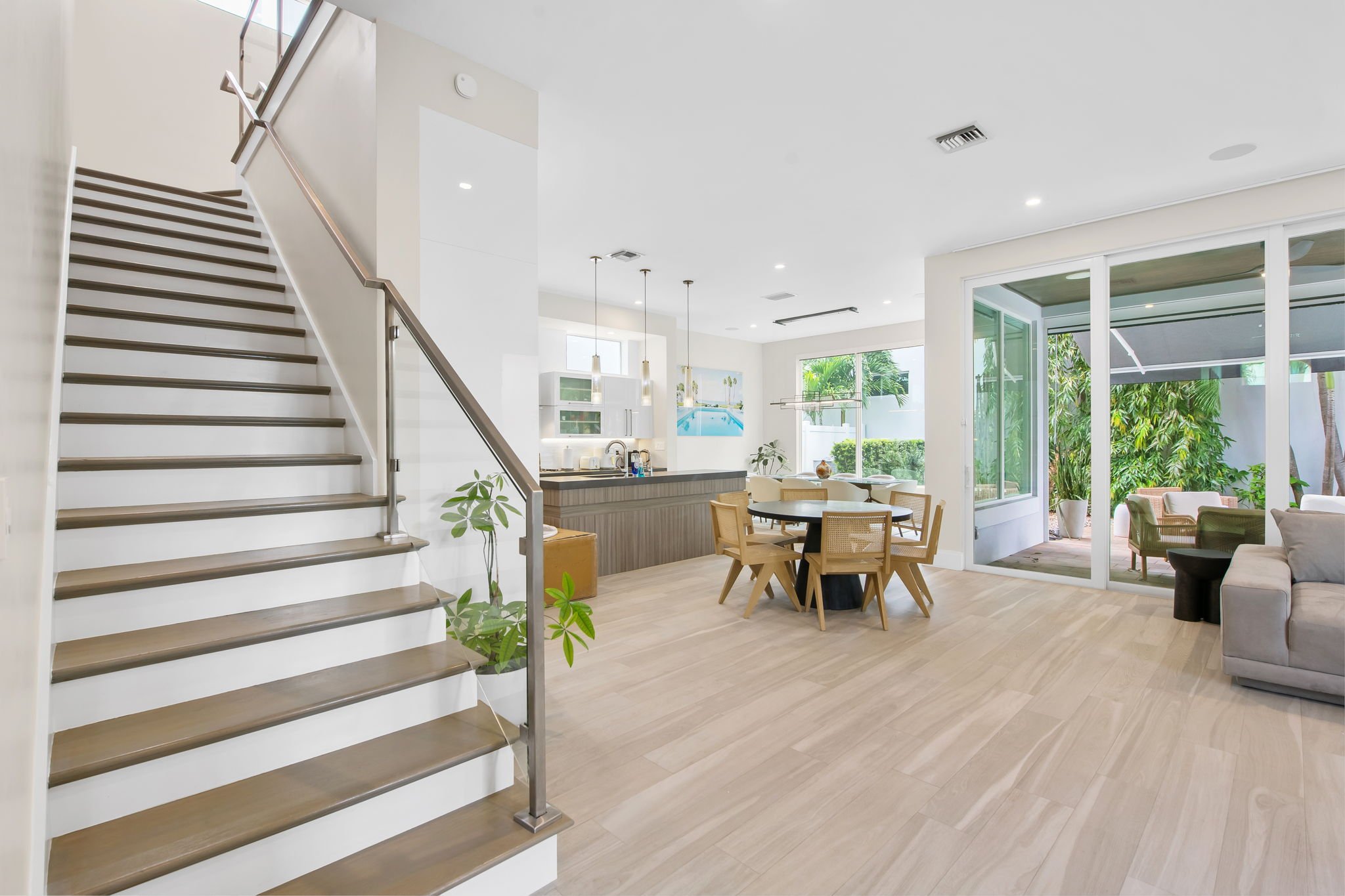 Bright open-concept living space with a staircase, kitchen, and dining area, featuring large sliding glass doors opening to an outdoor patio with greenery.