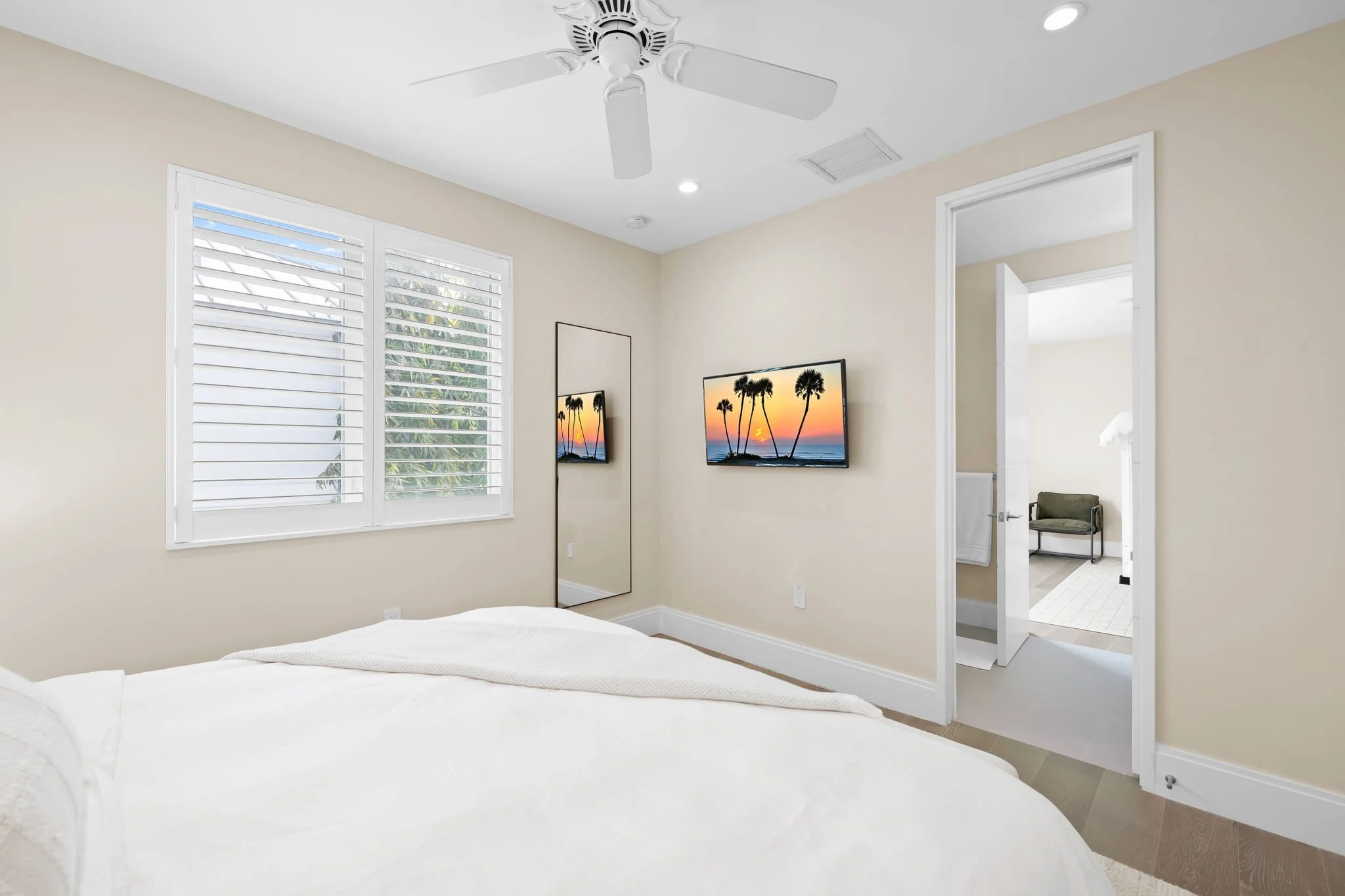 A bedroom with a ceiling fan, large window with shutters, two wall-mounted TVs showing a sunset with palm trees, an open doorway leading to a bathroom, a chair, and a bed with white bedding.