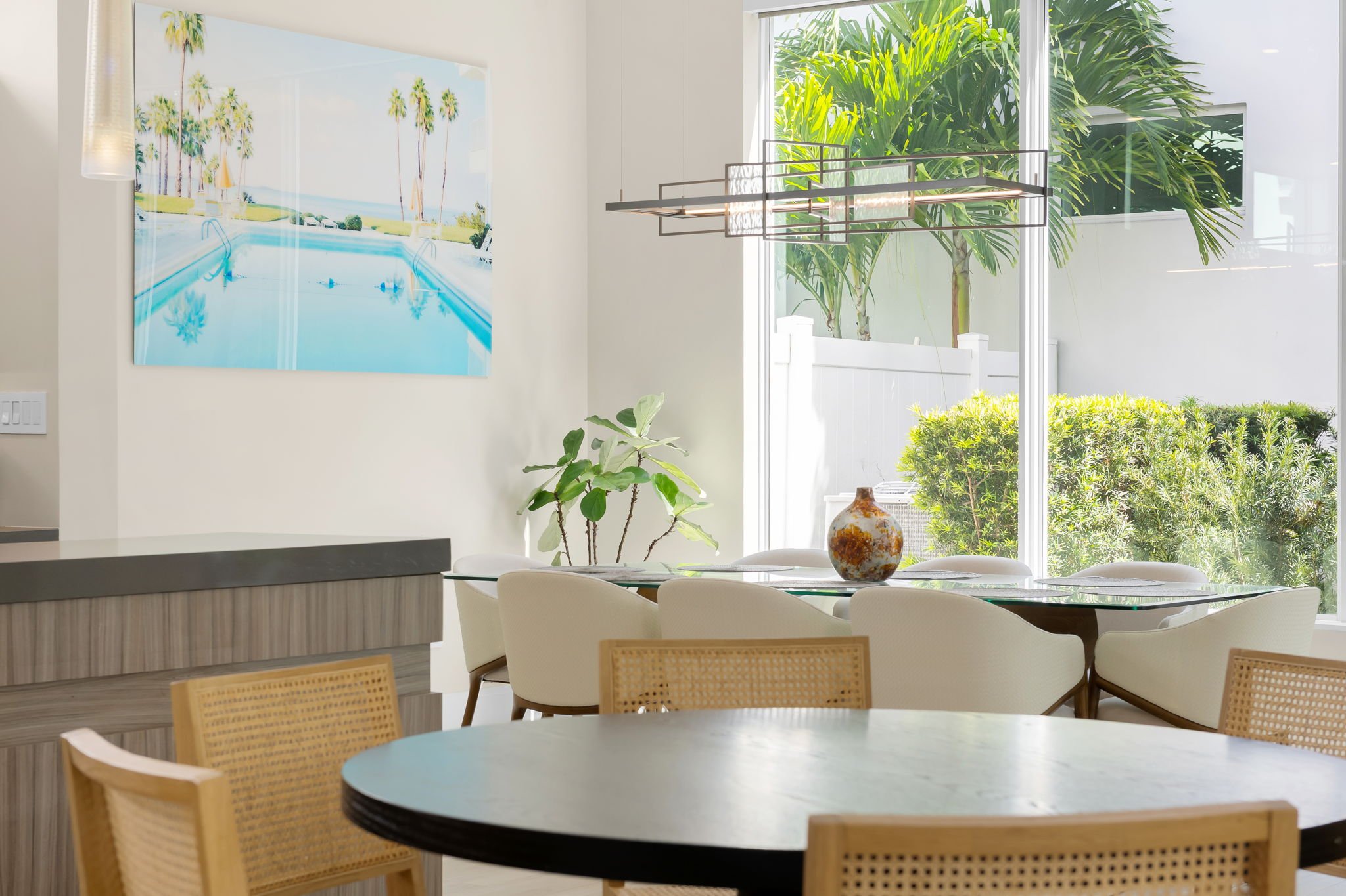 Modern dining area with white chairs around a round table, large window with view of greenery, large wall art of palm trees over a pool, and a contemporary chandelier.