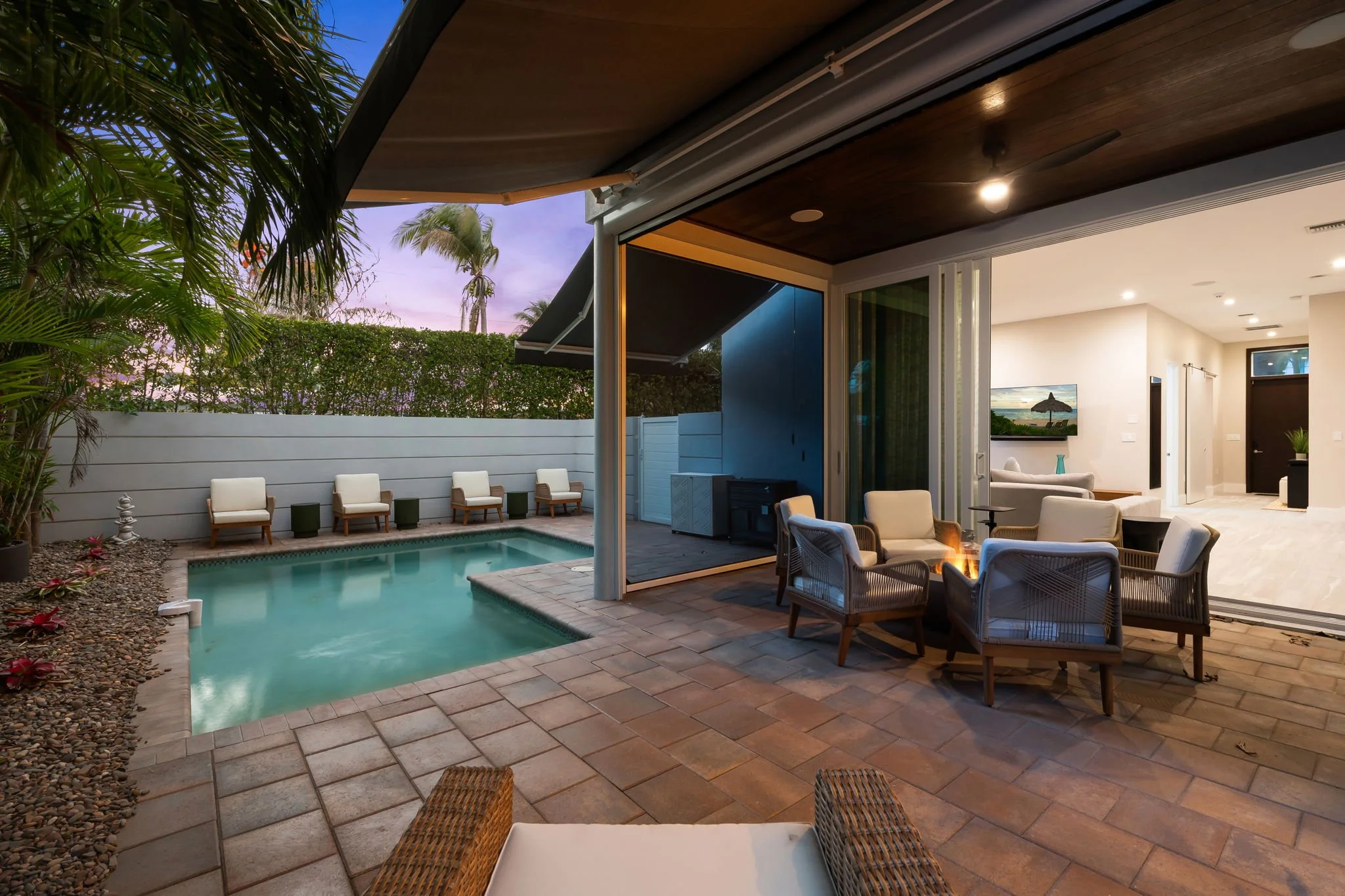 A luxurious backyard with a private swimming pool, outdoor seating, and a covered patio area inside a modern home, with a view of a sunset sky and palm trees in the background.