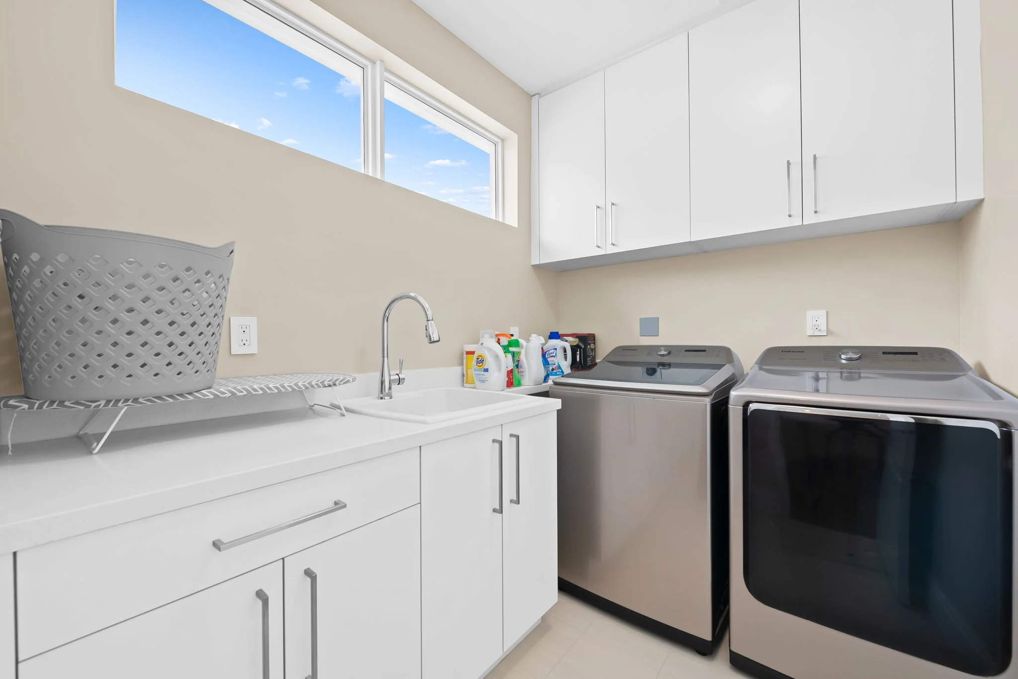 A laundry room with white cabinets, a sink, and a washer and dryer. Various laundry supplies are on the counter, and there is a window with a blue sky outside.