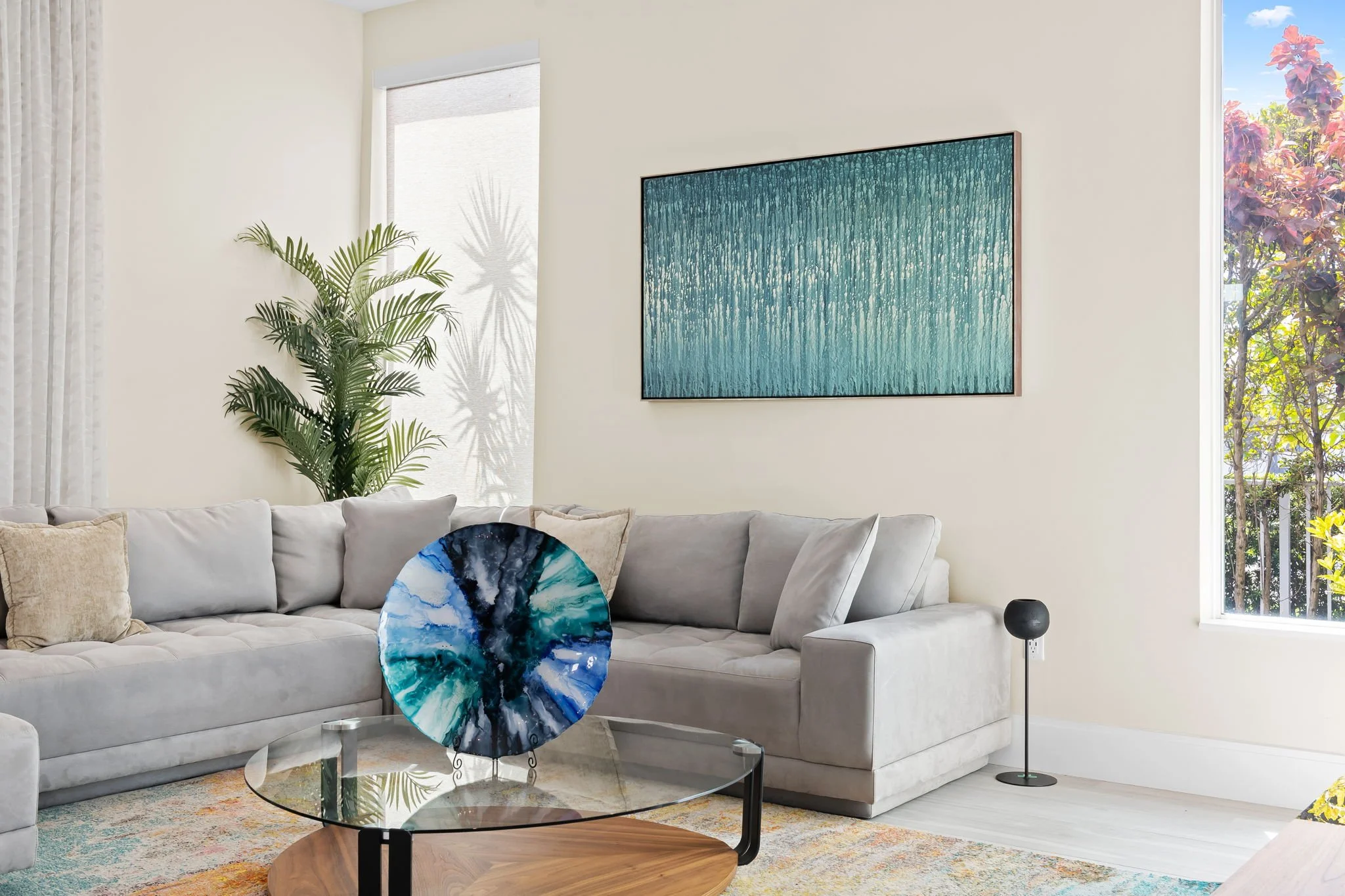 Living room with a beige sectional sofa, a glass coffee table, an abstract art piece on the wall, a potted plant, and a window with natural light.