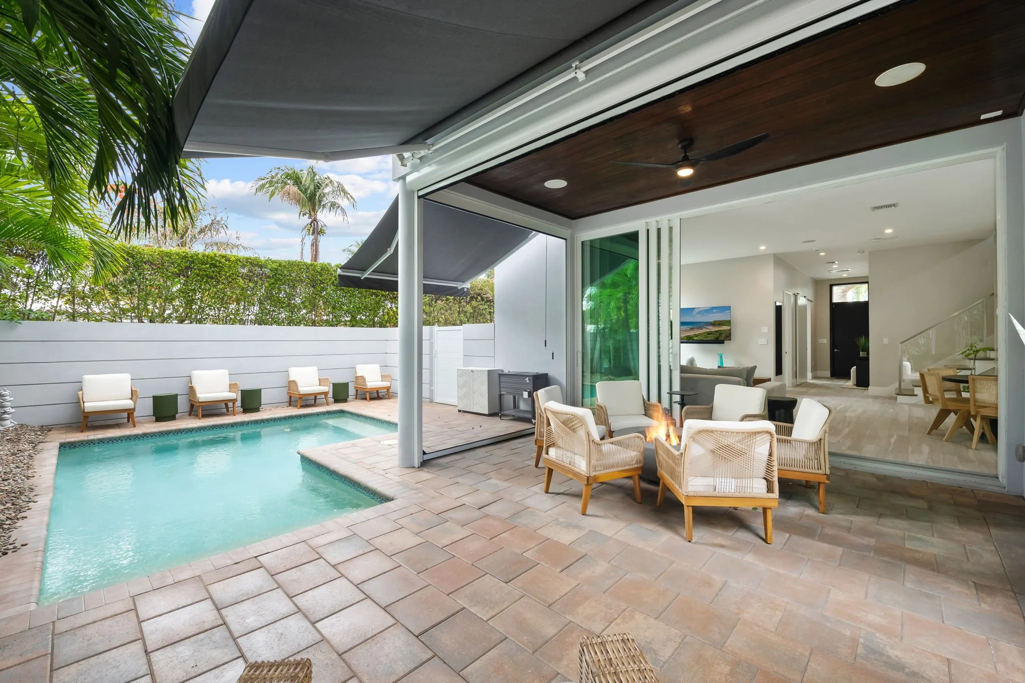 A view of an indoor-outdoor living space with a swimming pool, beige outdoor chairs, a gas fire pit, and a wall of greenery. The area opens to a modern interior with a TV, staircase, and large windows.