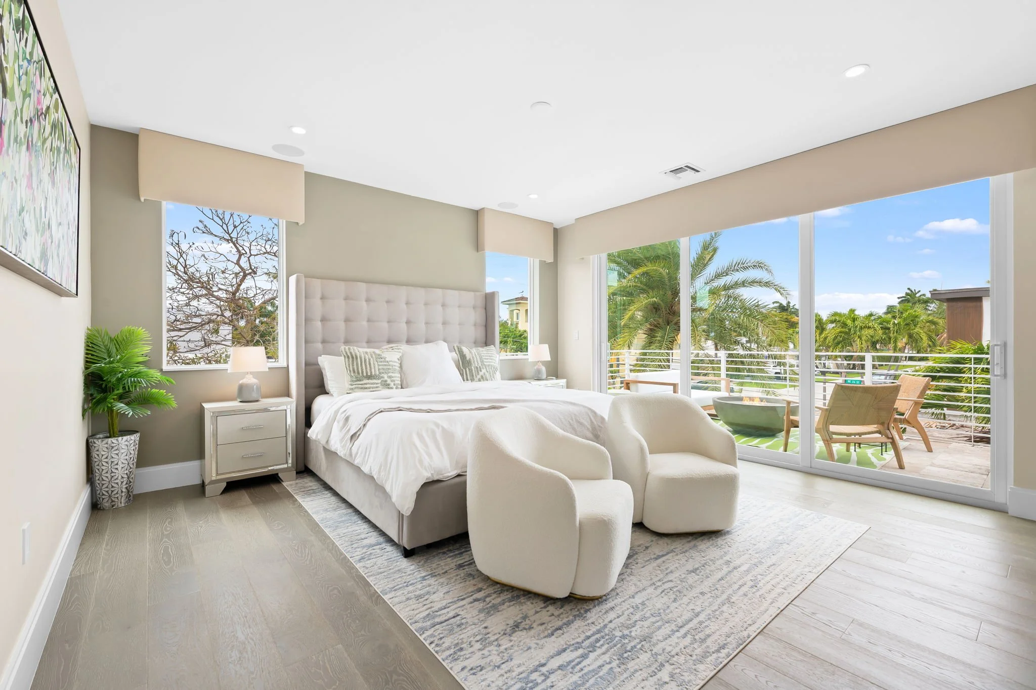 A bright bedroom with large windows showing a balcony and palm trees outside. The room features a white bed with a beige tufted headboard, white and patterned pillows, two nightstands with lamps, white armchairs, a potted plant, and abstract artwork 