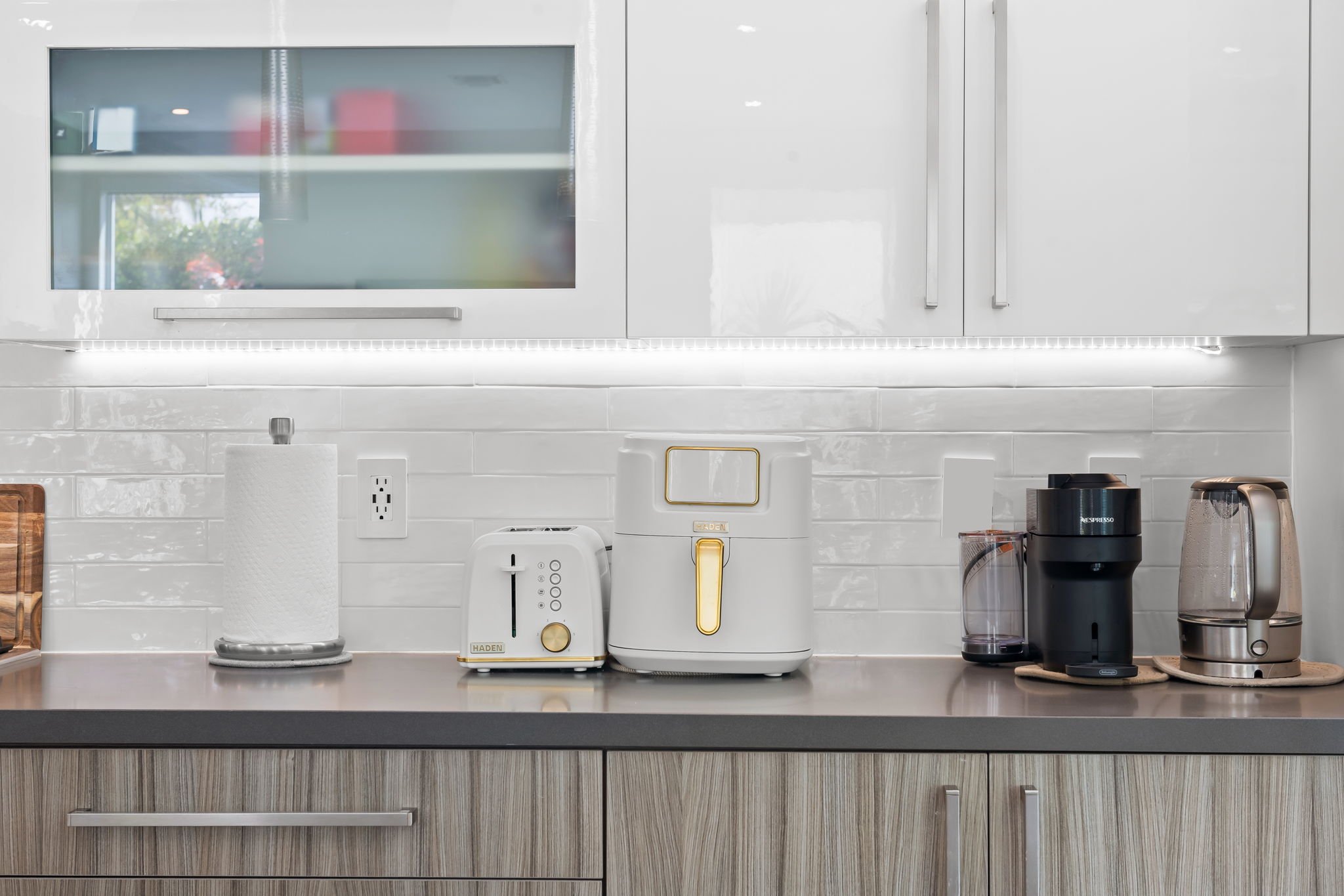 Kitchen countertop with small appliances including a white toaster, a white bread maker, and coffee machines, set against white cabinets and a tiled backsplash.