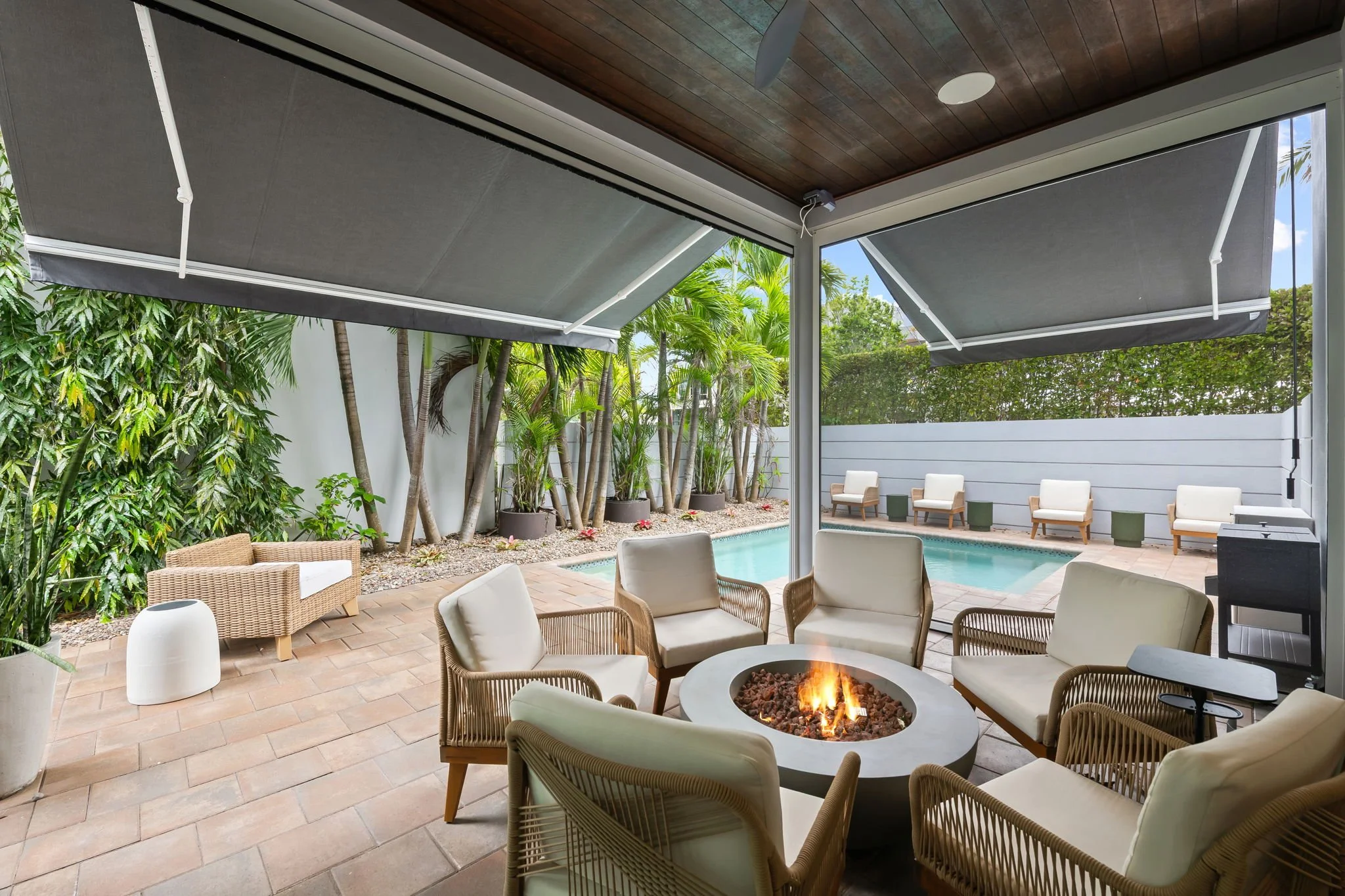 Covered outdoor patio area with lounge chairs, a firepit, lush greenery, and a pool in the background.
