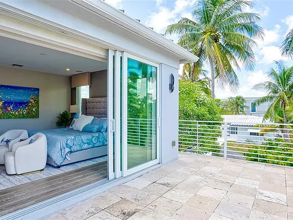View of a bedroom with a sliding glass door open to a balcony with tropical palm trees and a modern cityscape in the background.
