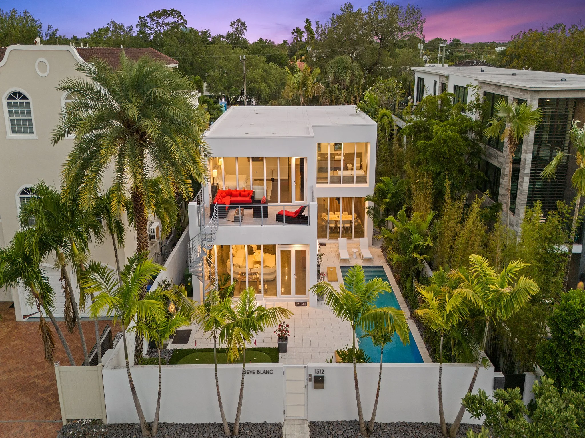 A modern white townhouse with large windows and a rooftop terrace, surrounded by lush green palm trees and vegetation, during dusk with a purple sky.