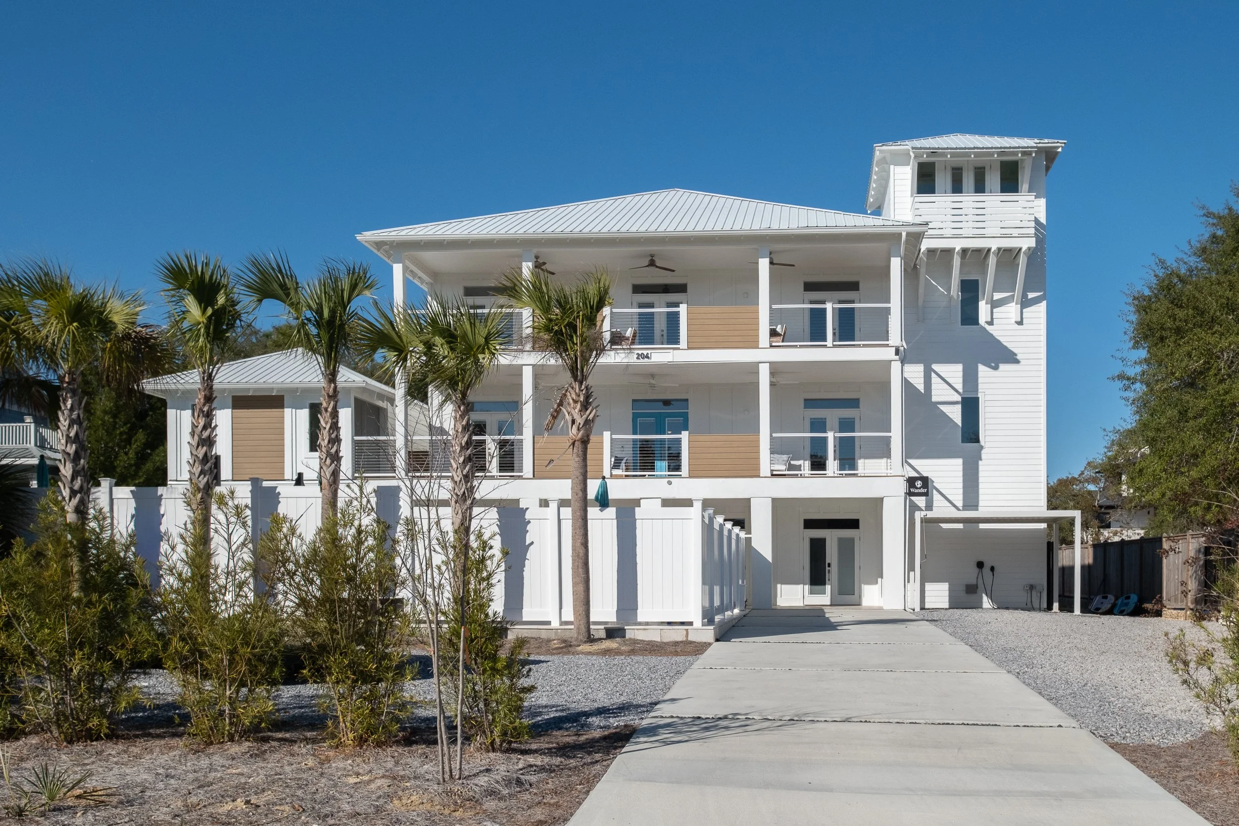 A white multi-story house with a metal roof, balconies, and a small tower on the right side. The house is surrounded by palm trees and a gravel yard with a concrete walkway leading to the entrance under a clear blue sky.