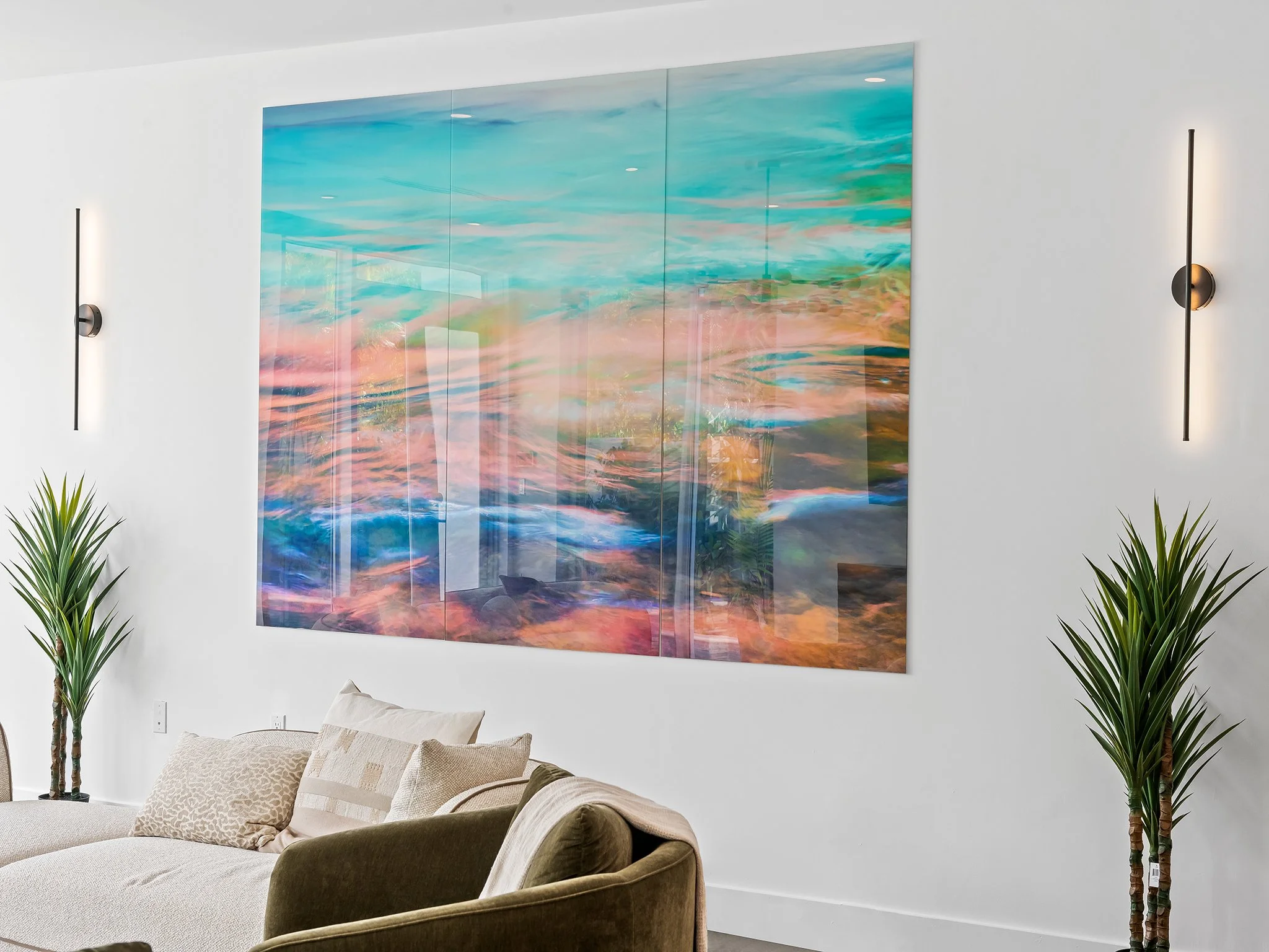 Living room with a large, abstract ocean-themed wall art, beige couch with pillows, and tall green plants flanking the artwork, illuminated by modern wall sconces.