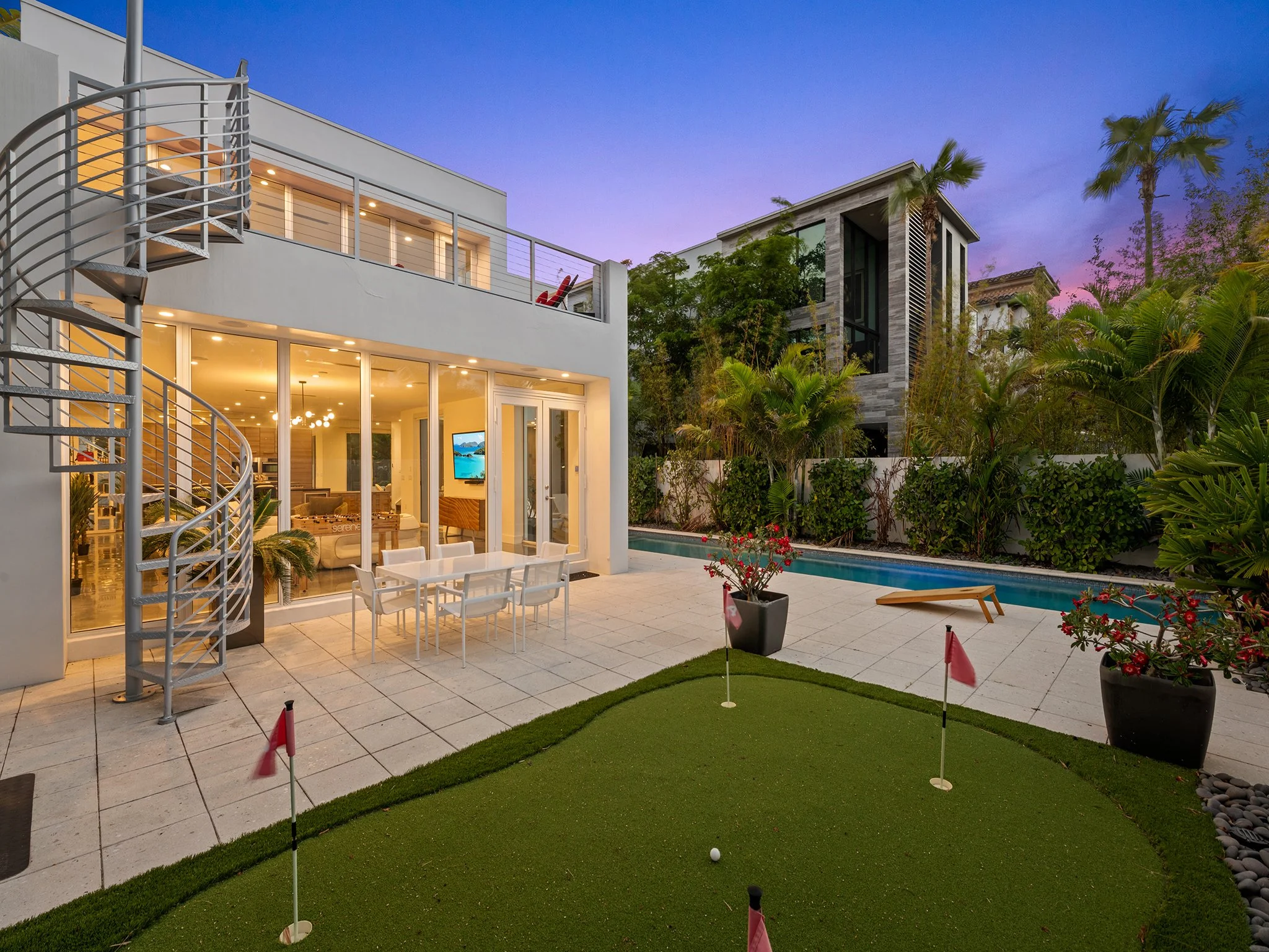 Modern backyard with a golf putting green, a pool, patio furniture, and two multi-story houses surrounded by trees and palm trees at dusk.