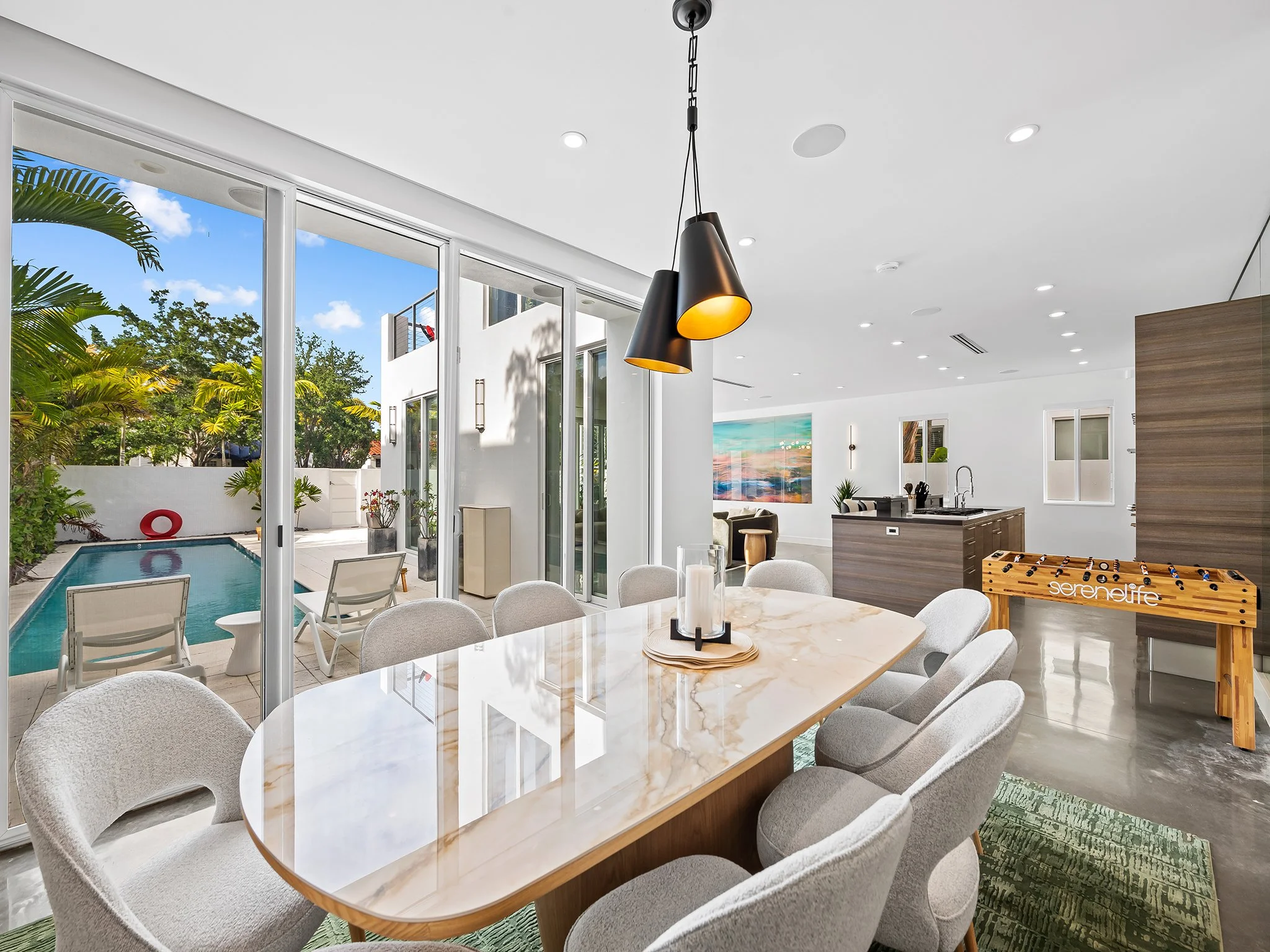 Modern indoor living room with large sliding glass doors leading to an outdoor pool area, white walls, a hanging pendant light, a marble dining table with chairs, a foosball table, and artwork on the wall.