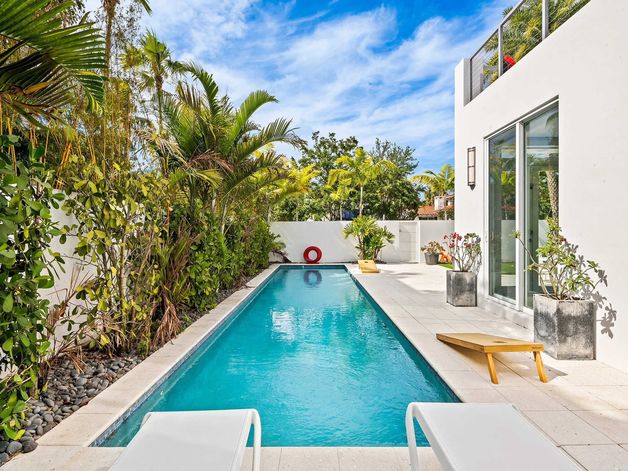 A backyard with a rectangular swimming pool, surrounded by potted plants and tropical trees, with a modern white house on the right and a blue sky with scattered clouds overhead.