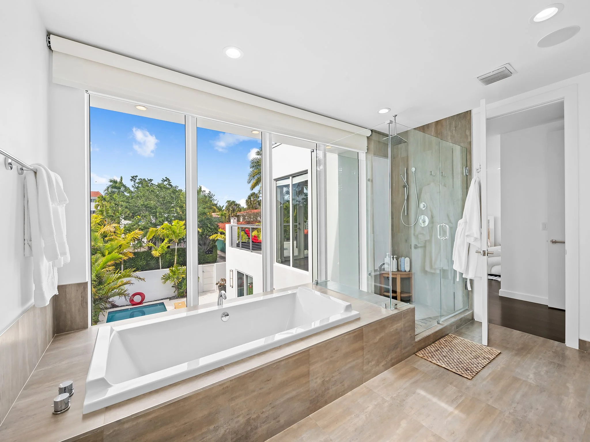 Modern bathroom with large window showing tropical trees, walk-in shower with glass enclosure, and a bathtub with a view outside.