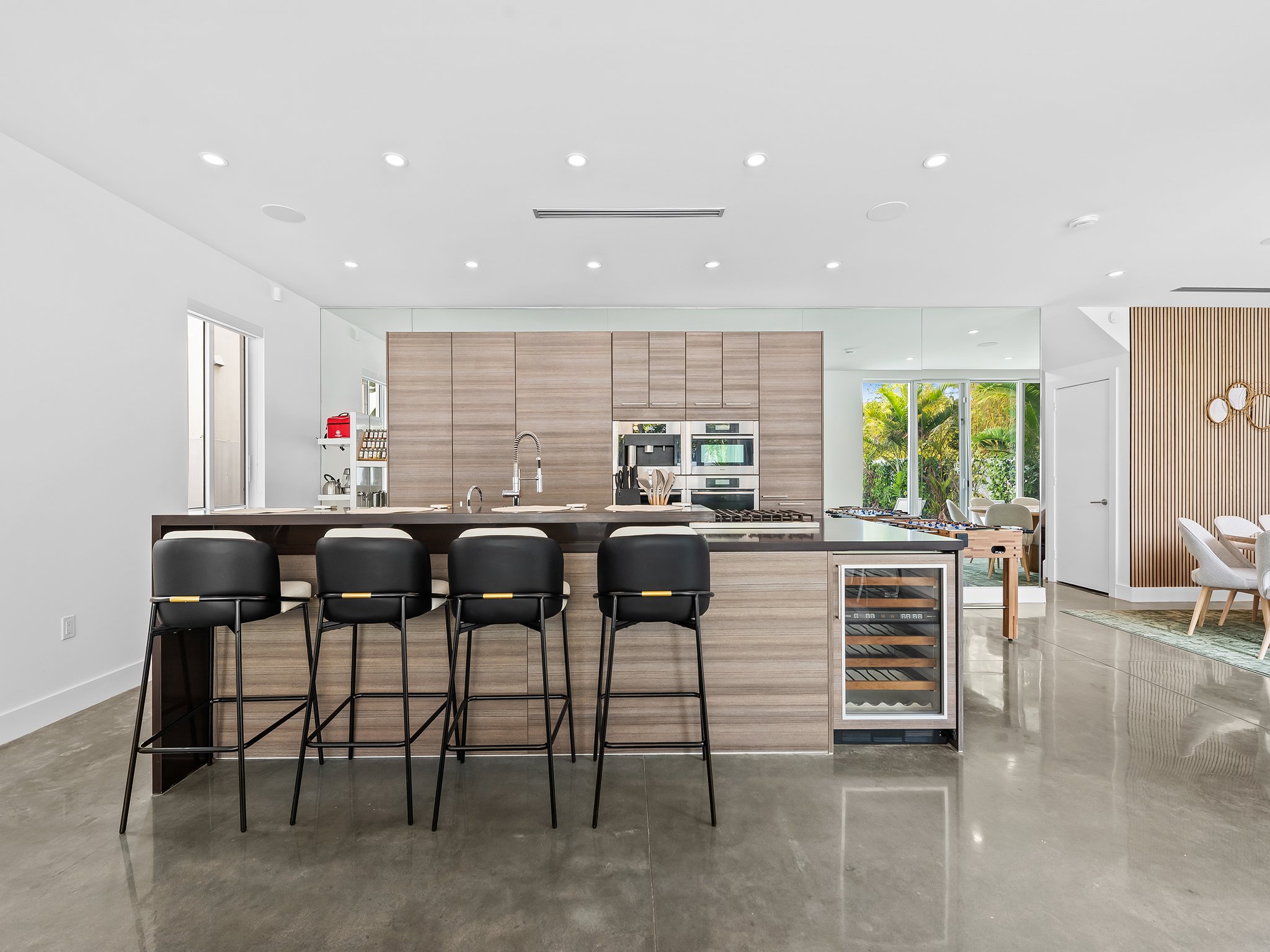 Modern kitchen with a large island, black chairs, wooden cabinetry, stainless steel appliances, and a view of greenery through large windows.