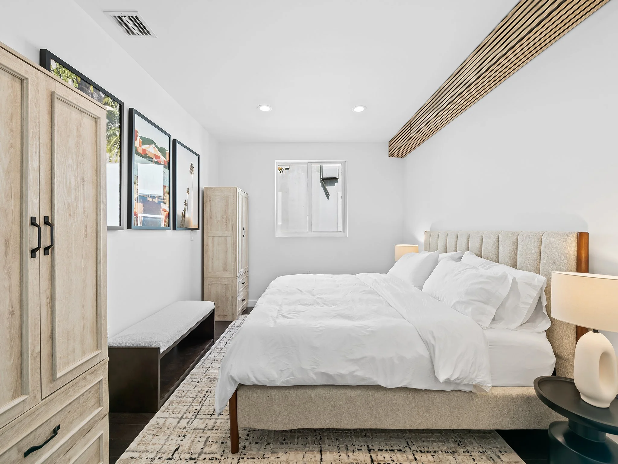 A modern bedroom with white walls, a bed with white linens, wooden furniture, framed artwork on the wall, a window, and a beige carpet.