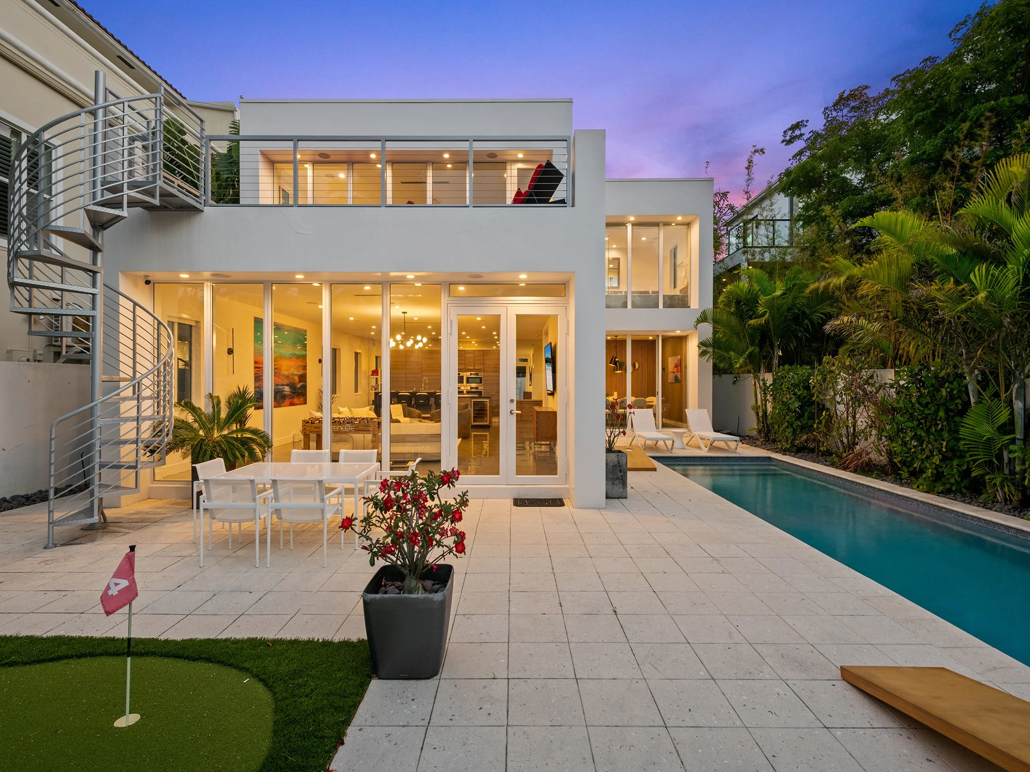 Modern backyard with a swimming pool, outdoor seating, potted plants, a small putting green, and a multi-story white house with large windows and balcony, surrounded by greenery at dusk.