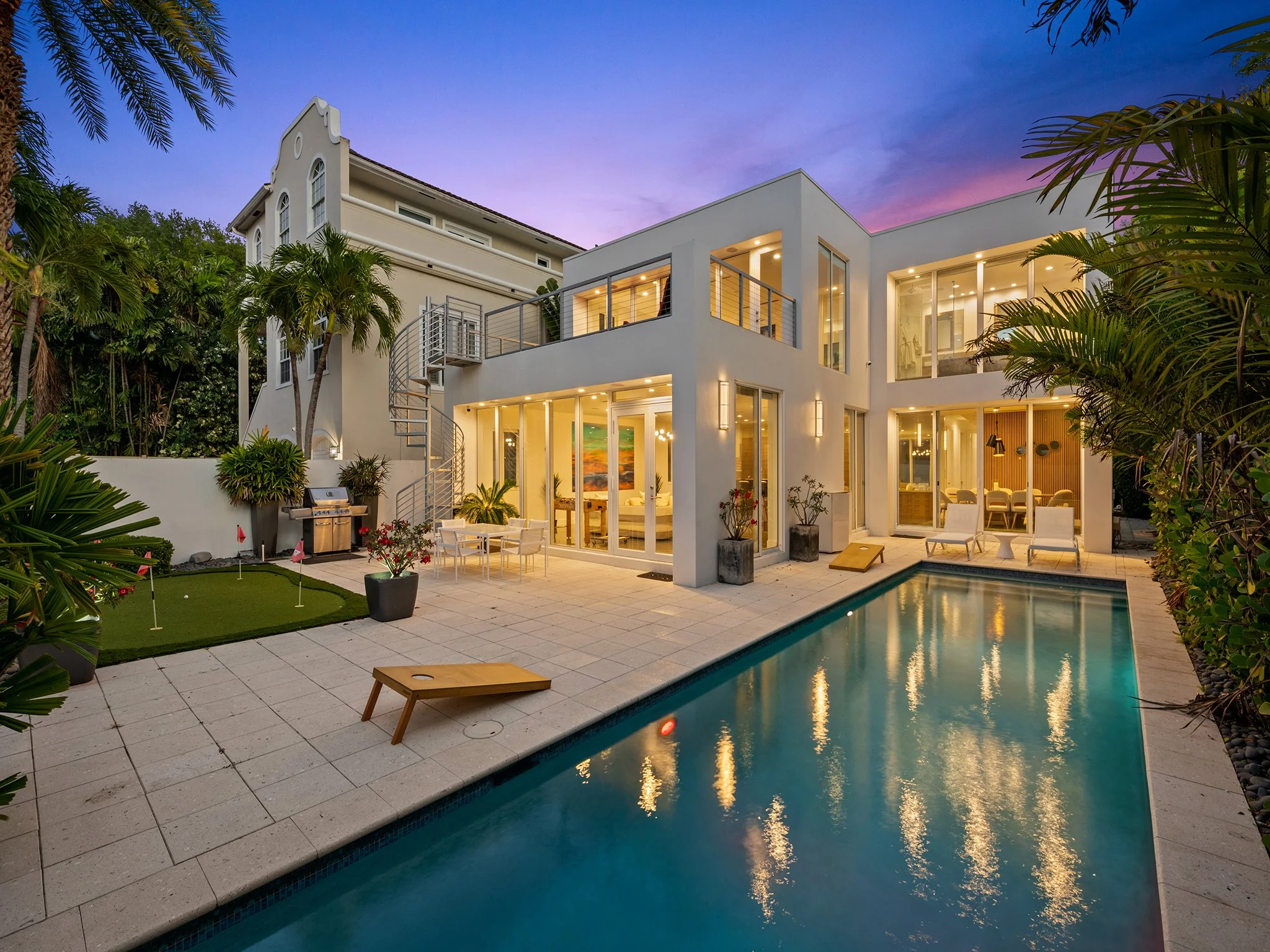 Modern white house with large glass windows and outdoor patio, overlooking a swimming pool and surrounded by lush green tropical plants during sunset.