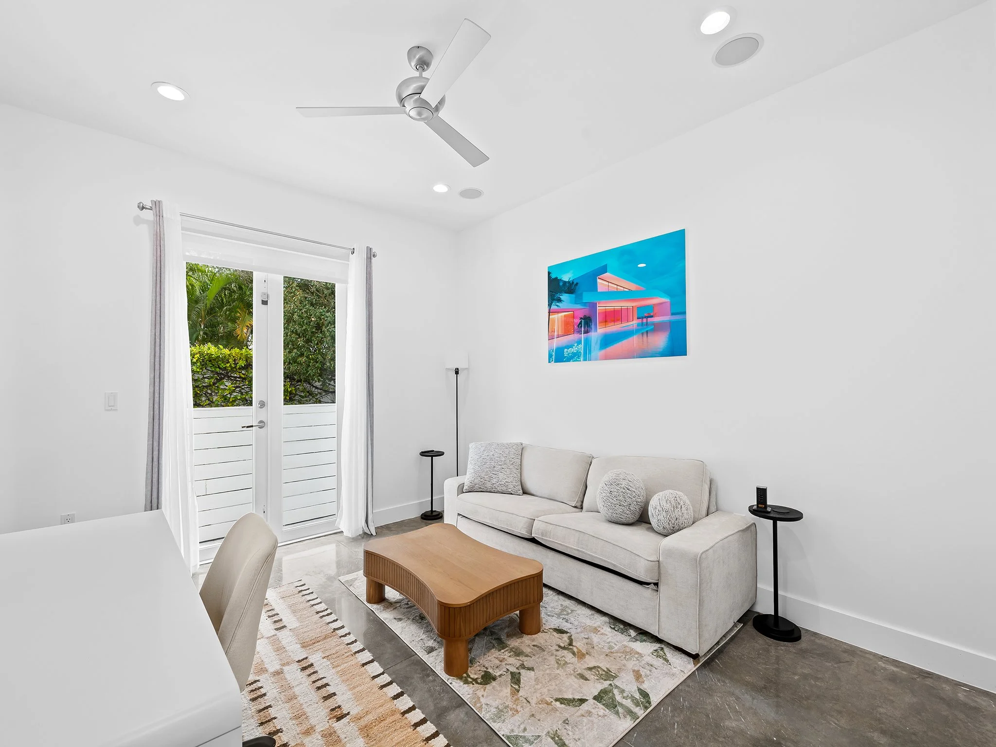 Living room with white walls, beige sofa, wooden coffee table, nearby patterned rug, large sliding glass door with white curtains, wall art of a modern house, ceiling fan, and windows showing greenery outside.