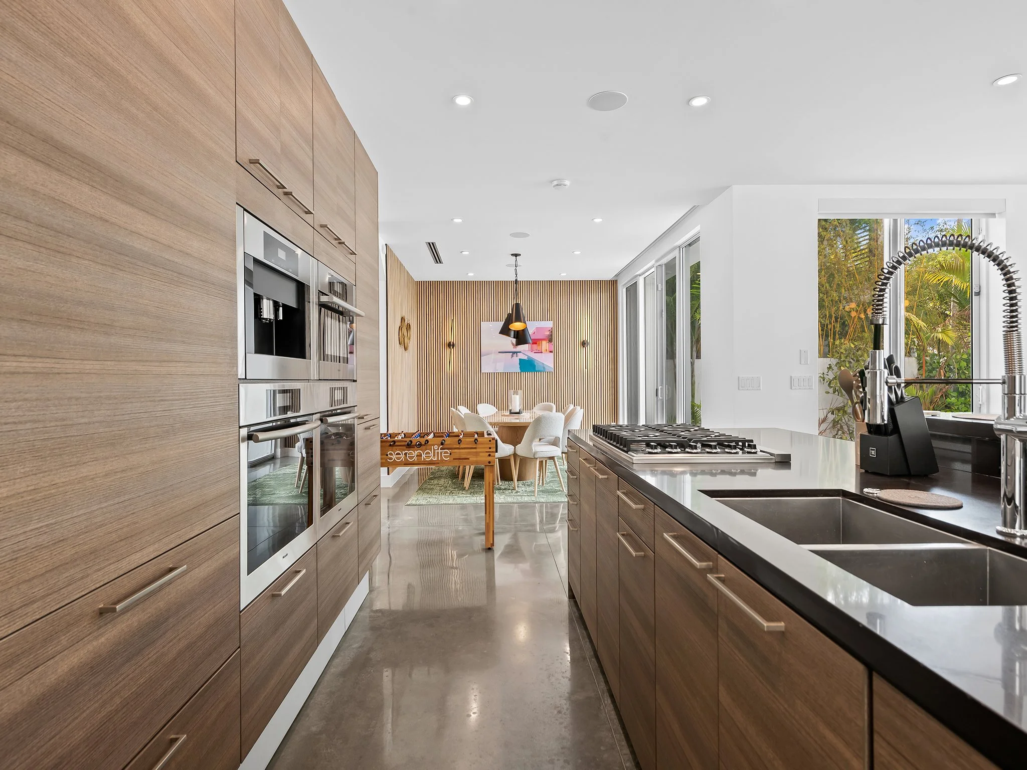 Modern kitchen with wooden cabinets, stainless steel appliances, black countertops, large window showing outdoor greenery, and dining area in the background.