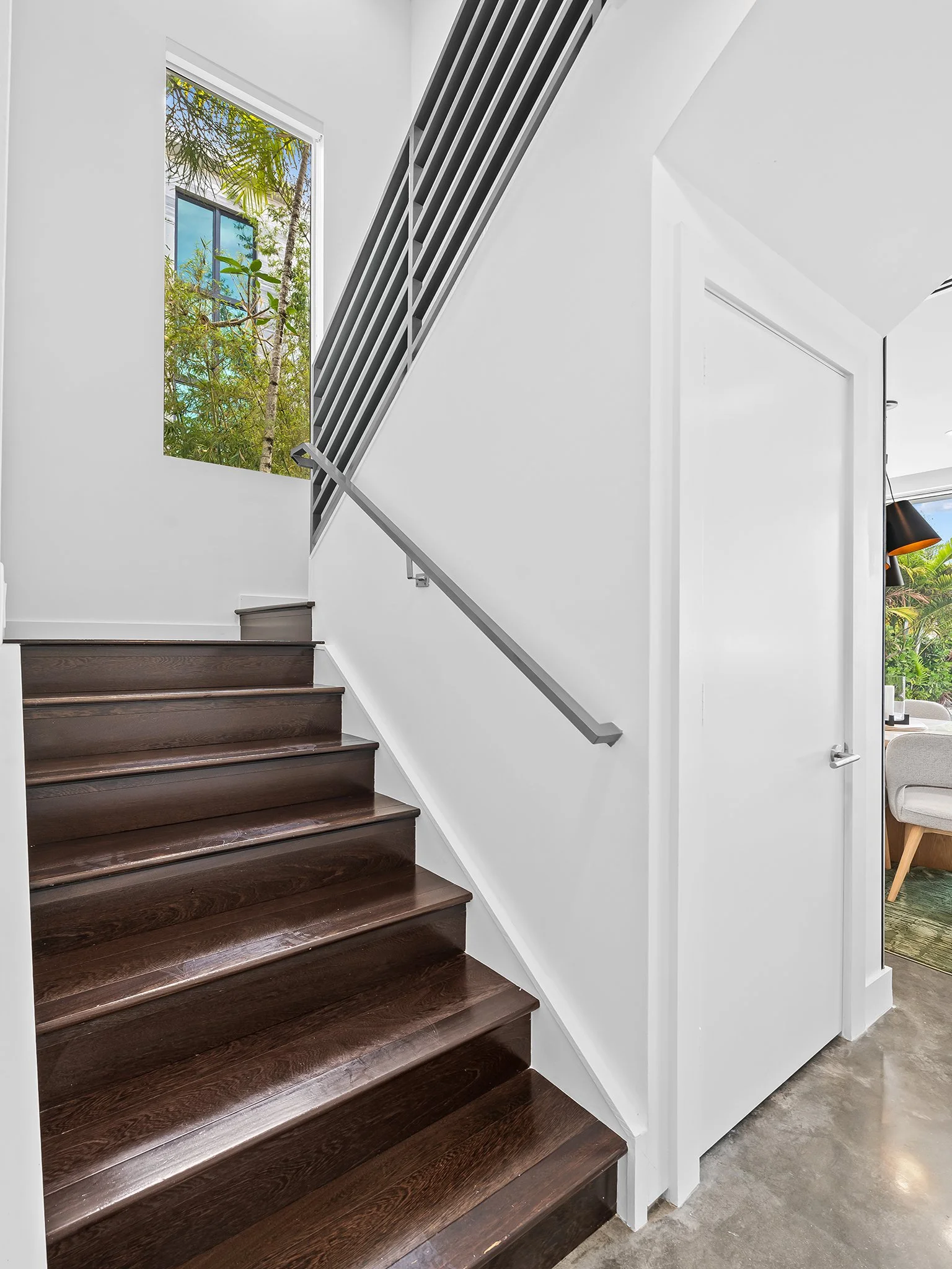Interior view of a modern staircase with dark wooden steps, a white wall, and a window showing greenery outside, in a brightly lit space.