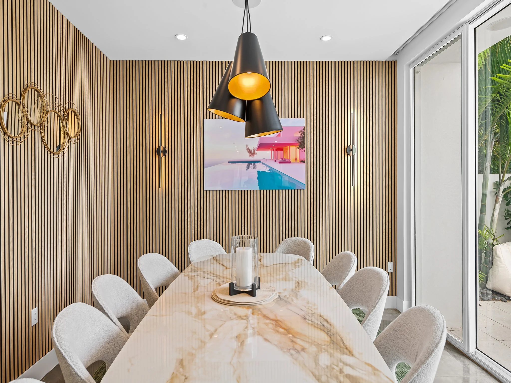 A dining room with a marble table surrounded by chairs, a wooden slatted wall, and a view of a pool area through sliding glass doors, with hanging ceiling lights.
