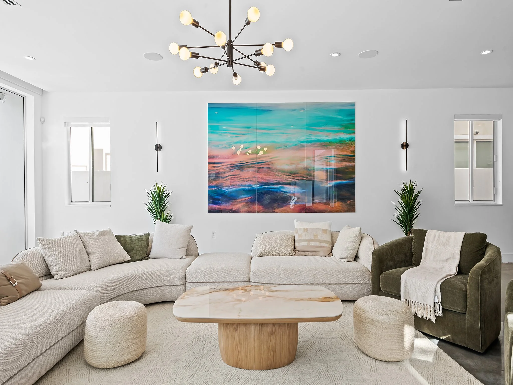 Modern living room with a white sectional sofa, decorative pillows, a wooden coffee table, a large colorful abstract painting on the wall, two green potted plants, a contemporary chandelier, and windows letting in natural light.