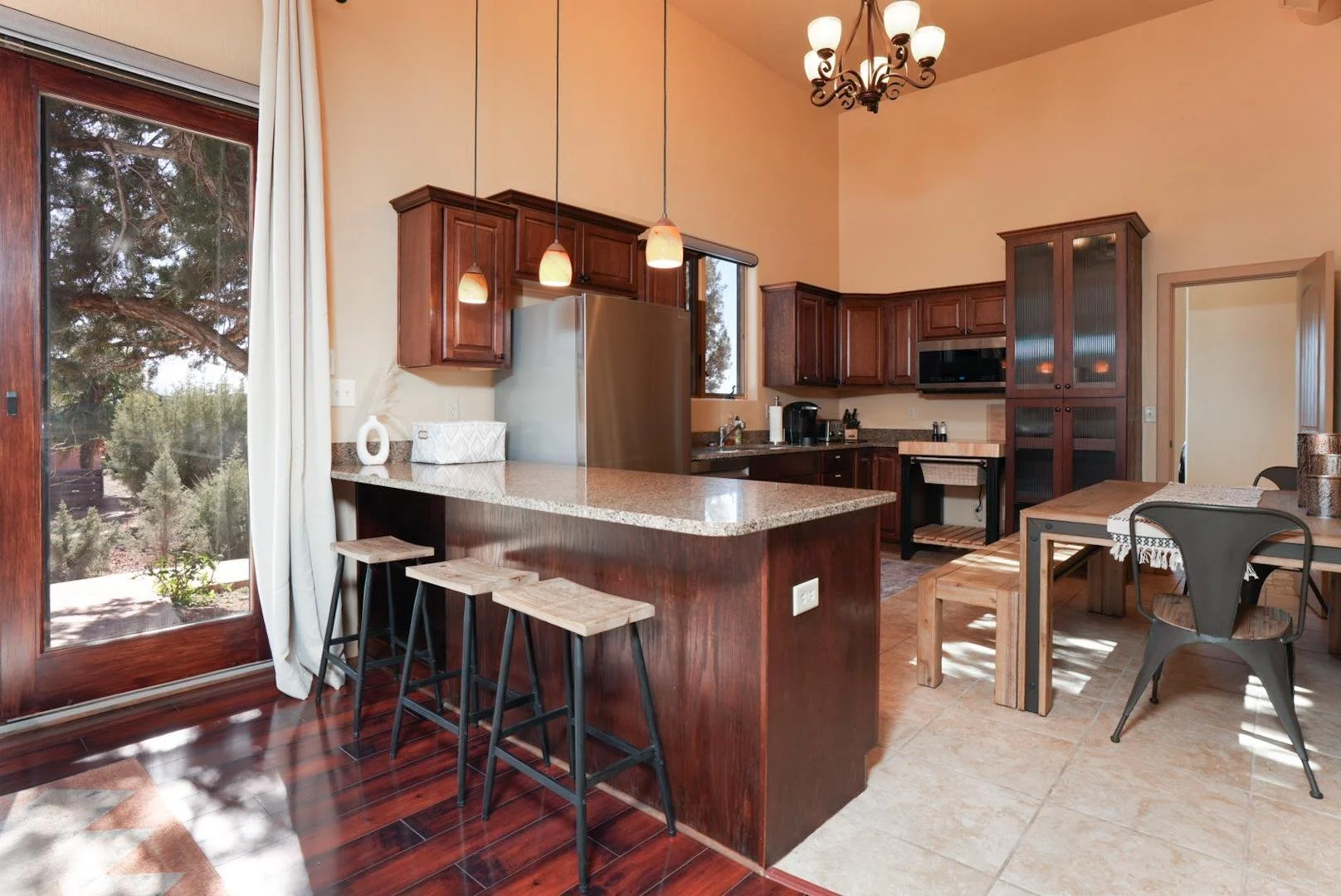 Kitchen with wooden cabinets, granite countertops, stainless steel refrigerator, pendant lights, a dining table with chairs, and a sliding glass door with white curtains leading outside.