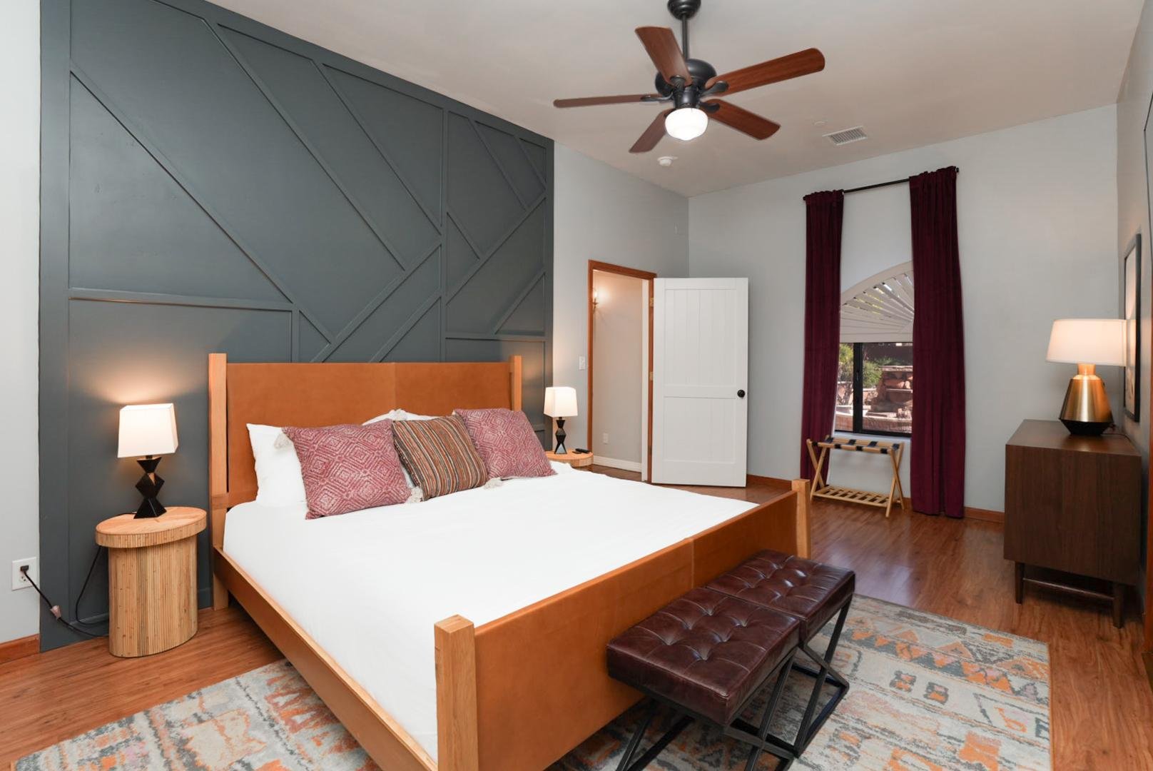 Modern bedroom with a wooden bed frame, quilted headboard, and multiple pillows. There is a dark wood bench at the foot of the bed, and side tables with lamps. The room has hardwood floors, patterned area rug, a window with dark red curtains, and a c
