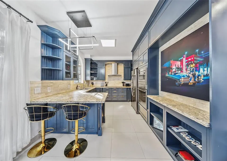 Modern kitchen with blue cabinetry, granite countertops, and a large flat-screen TV on the wall.