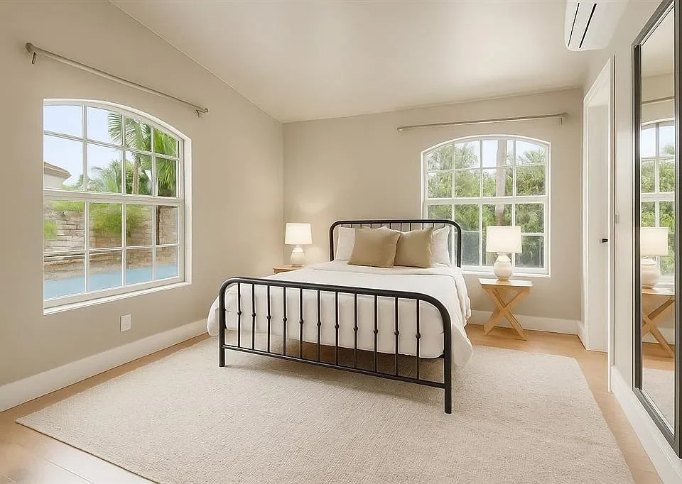 A bedroom with a bed, two bedside tables with lamps, two windows, and a mirrored closet door.
