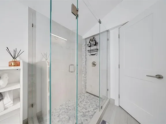 Modern bathroom with a glass shower enclosure, white storage shelf, and grey door.