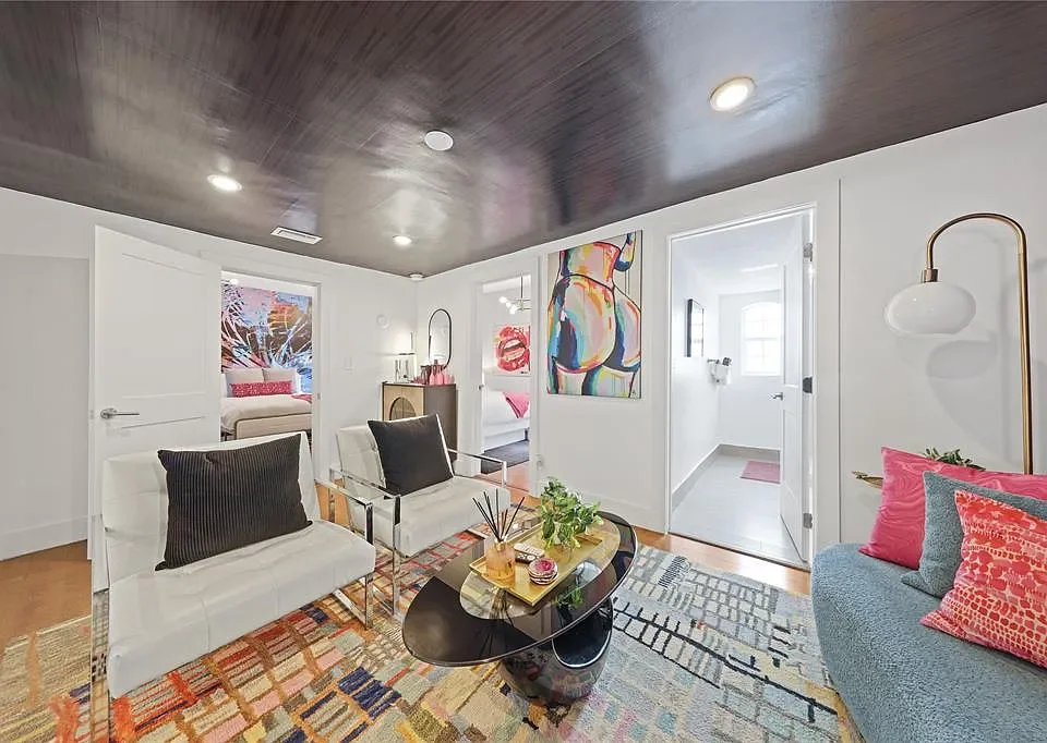Living room with white walls, dark wood ceiling, colorful abstract art, and modern furniture including white sofas with pink and black pillows, a black coffee table with decor, and an area rug. Two doorways lead to bedrooms or other rooms, with the b