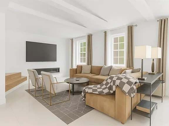 Living room with beige sofa, white armchairs, a round coffee table, a large flat-screen TV, a bookshelf, and multiple windows with beige curtains.