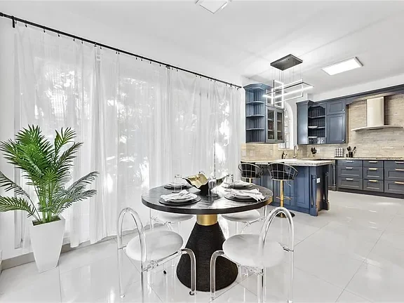 Modern kitchen and dining area with white curtains, a black round dining table set for four, transparent chairs, and a blue kitchen with open shelves and a light-colored backsplash.